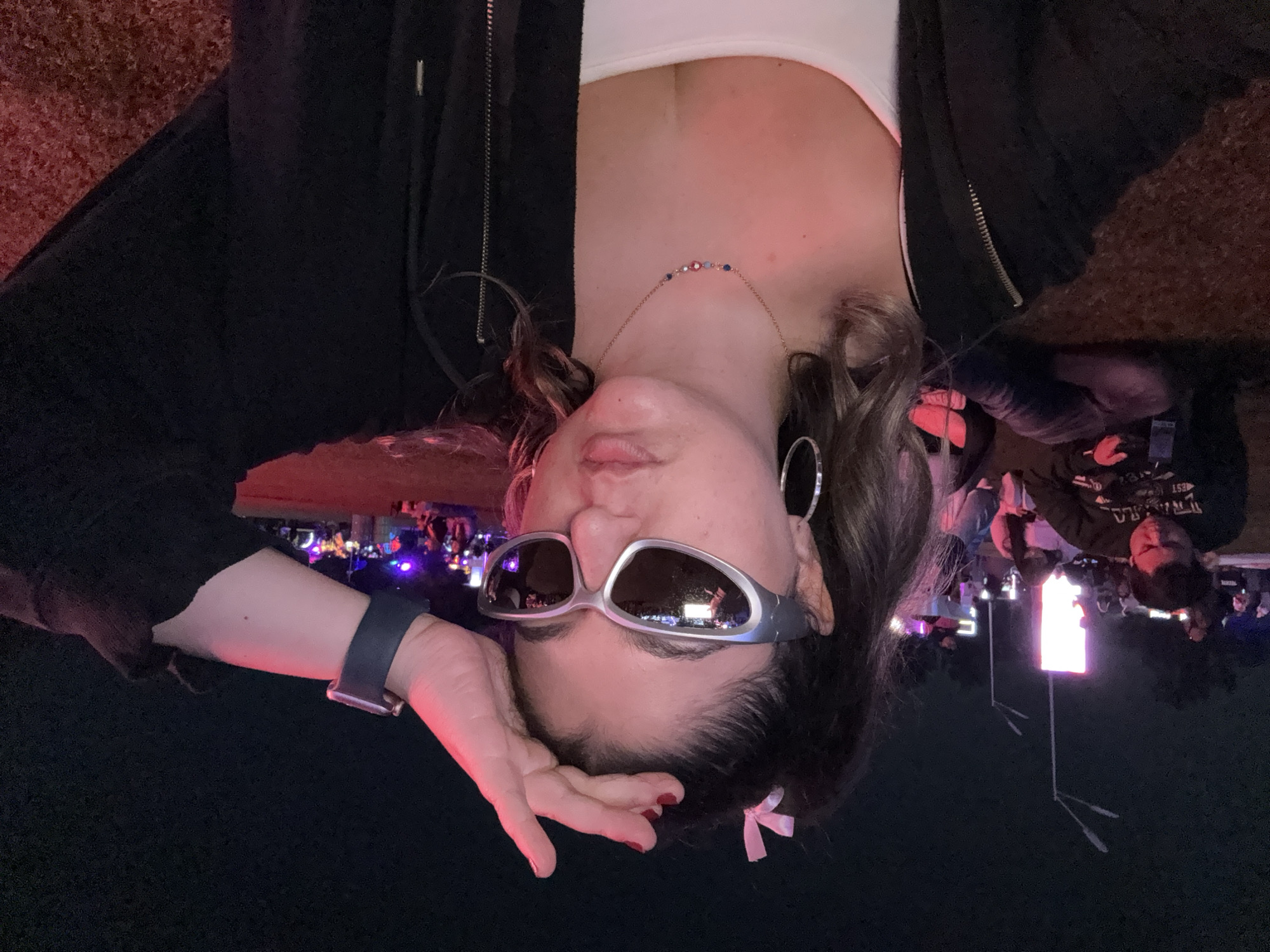 A woman is posing for a photo. She is wearing silver sunglasses, a white tank top, and a black zip-up hoodie. She has hoop earrings and a necklace. She is wearing an Apple Watch on her left wrist. Her hair is dark and curly, and there is a pink ribbon in her hair. The background is dark, but there are people and lights visible. It looks like she is at a festival or concert. It is not possible to determine the exact location within Mexico City.