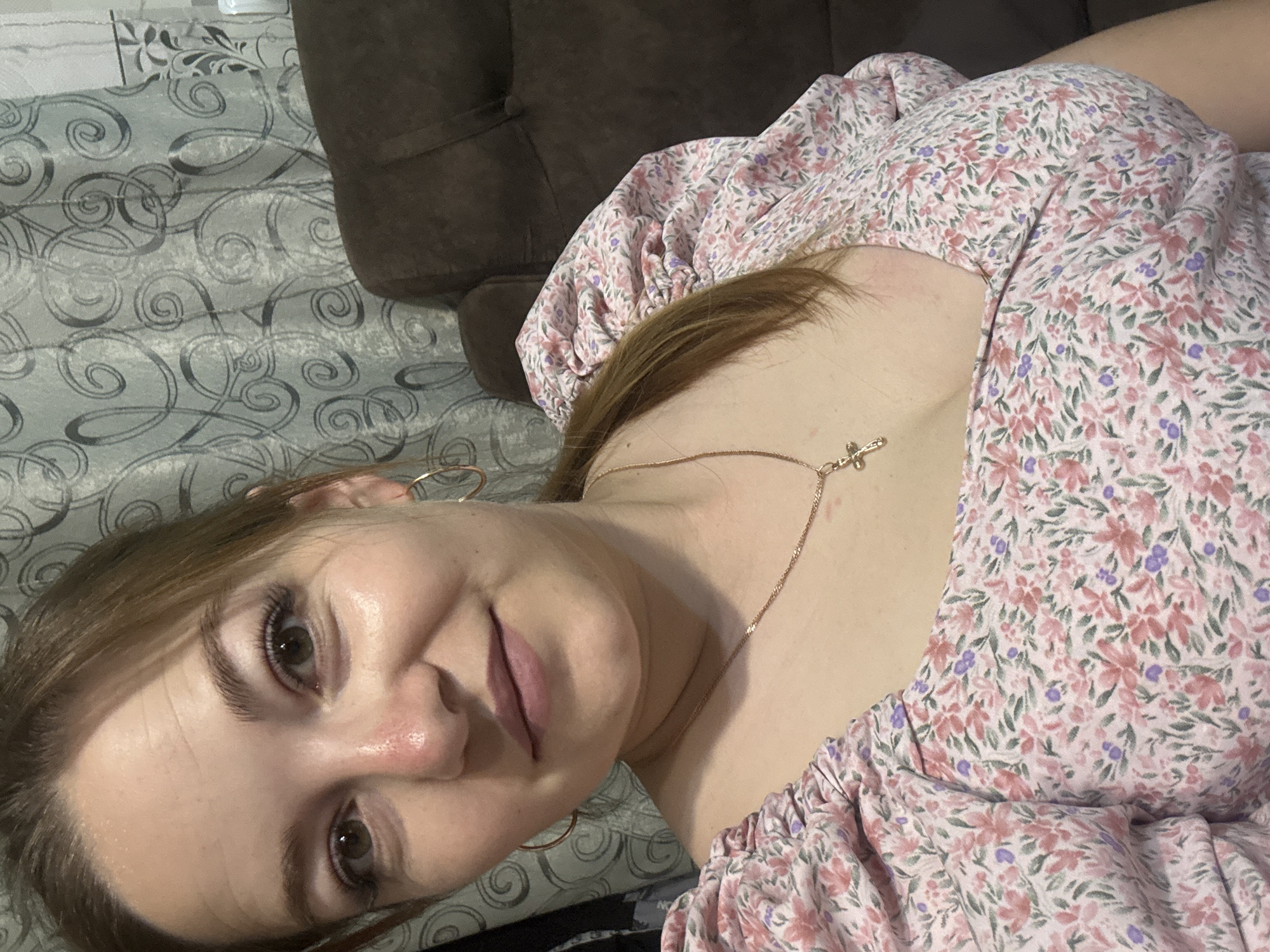 The image is a close-up, selfie-style photograph of a young woman with light brown hair and green eyes. She is wearing a pink floral print dress with puff sleeves and a gold necklace with a cross pendant. She is also wearing hoop earrings. The background consists of a patterned curtain and a dark brown upholstered chair. The scene appears to be indoors, possibly in a residential setting, in the city of Zubov, Russia.