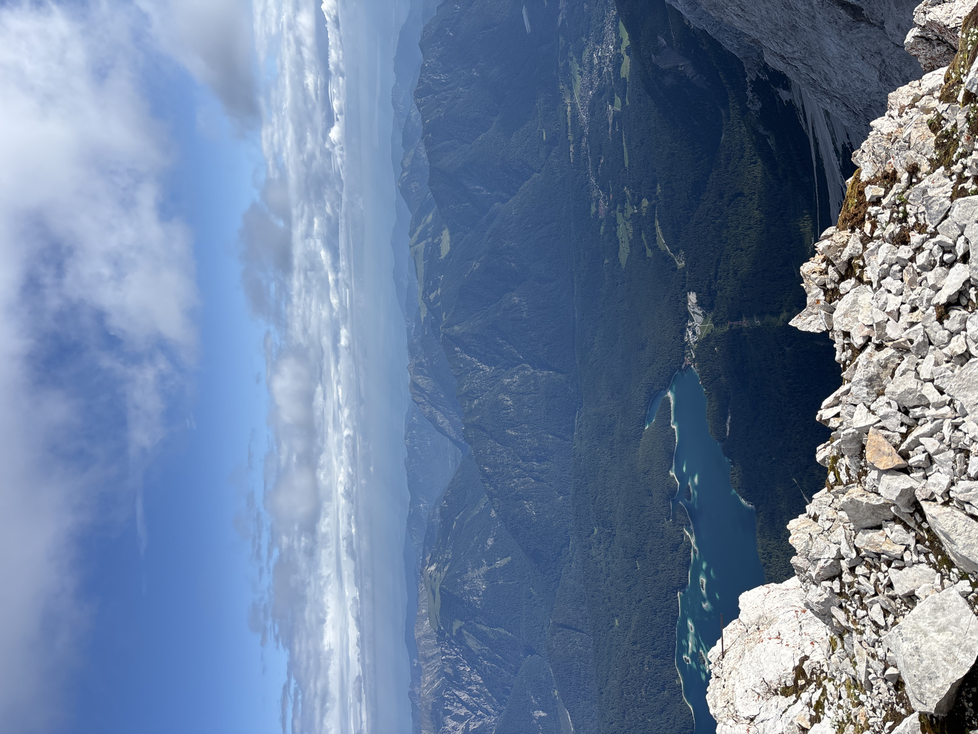 The image depicts a panoramic view of a mountainous landscape, with a bright blue sky dotted with white clouds. In the foreground, a rocky, grey terrain with sharp, irregular stones dominates the lower part of the frame. A vibrant turquoise lake is visible in the middle ground, surrounded by dark green, densely forested mountains. Further in the background, layers of progressively lighter blue mountains recede into the distance. A small town or village with scattered buildings and green fields can be seen nestled in the valley. The overall scene suggests a high-altitude vantage point, offering a vast and natural vista. The scene is characteristic of the Alps, and given the mention of Ehrwald, Austria, it is likely a view from a mountain peak in or near the Ehrwald region. No people or specific actions are discernible in the image.