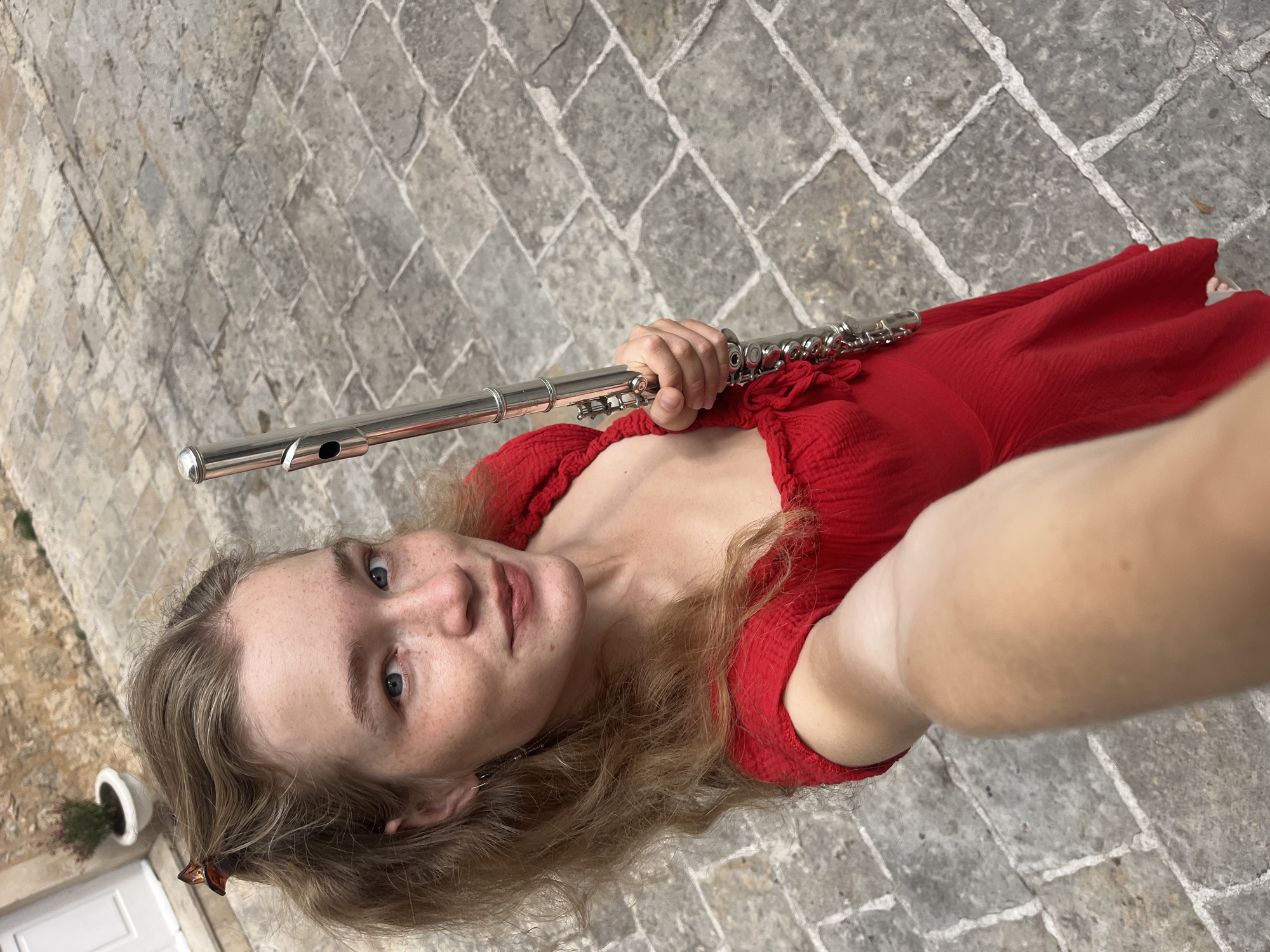 A selfie captures a woman in a red dress holding a flute, taken at an angle looking down. The woman has fair skin, freckles, blue eyes, and light brown hair. She is wearing a red, peasant-style dress. In her right hand, she holds a silver flute. The background is a paved stone surface. In the lower left corner of the image, a white door and a potted plant are partially visible. The setting is likely in the old city of Budva, Montenegro, given the style of paving stones and architecture.