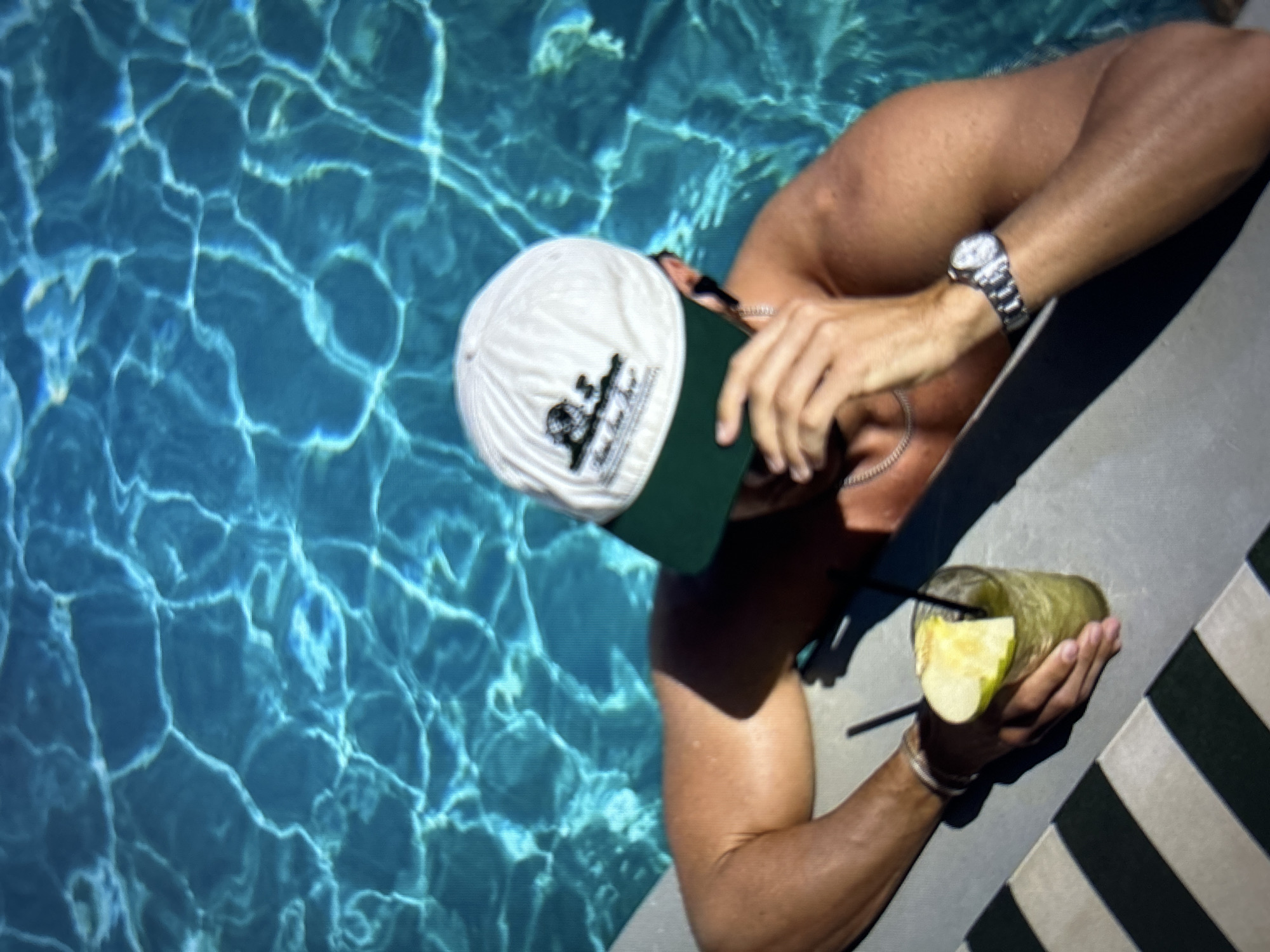 A man wearing a white baseball cap with a green brim is partially submerged in a swimming pool. The cap has a logo on the front. He is wearing a silver chain necklace and a silver watch on his left wrist. He is holding a glass with a green beverage and a slice of apple. The water in the pool is turquoise with light patterns reflecting on the surface. The man is leaning on the edge of the pool, which has a striped pattern. The scene appears to be in Burgas, Bulgaria, possibly at a hotel or private residence with a pool.