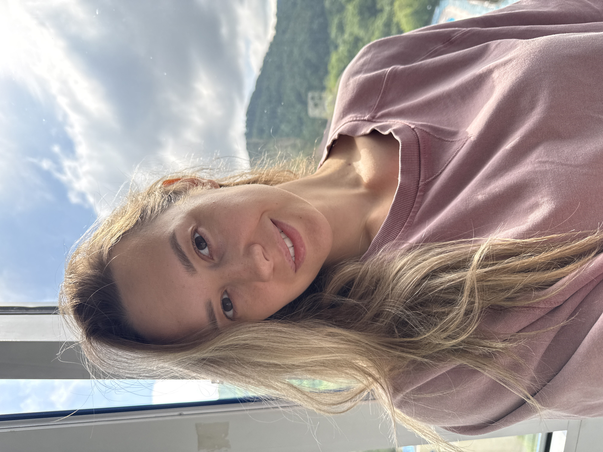 A medium shot photo shows a woman with blonde hair and brown eyes. She is wearing a pink crew neck sweatshirt and smiling at the camera. The background is blurred and shows a mountainous landscape with trees and buildings. The location is Nebug, Russia.