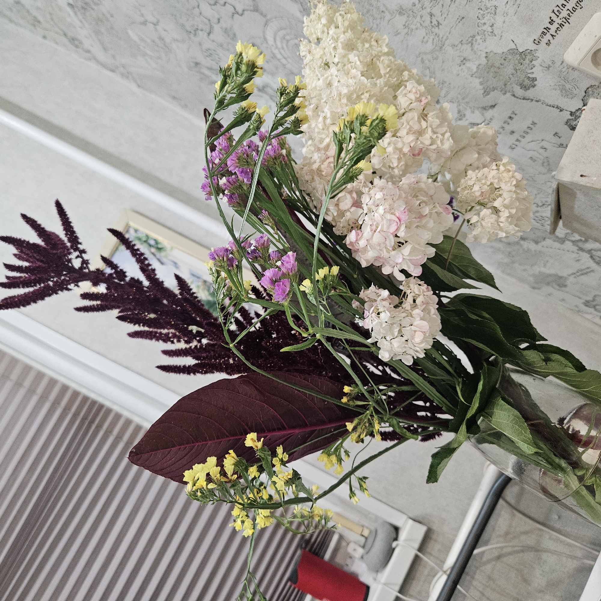 A bouquet of flowers is displayed in a clear glass vase. The bouquet includes deep purple amaranth, delicate pink and white hydrangeas, small yellow flowers, and thin purple statice. A large, dark red leaf is prominent on the left side of the arrangement. The background features a window with blinds, a textured gray wall with a map-like pattern, and a portion of a framed picture. Objects visible in the background include a red cylindrical object, a grey cylindrical object, and electrical outlets on the wall. The scene appears to be indoors, likely in a domestic setting. There is no indication of the city of Chelyabinsk, Russia, in the image or its metadata.