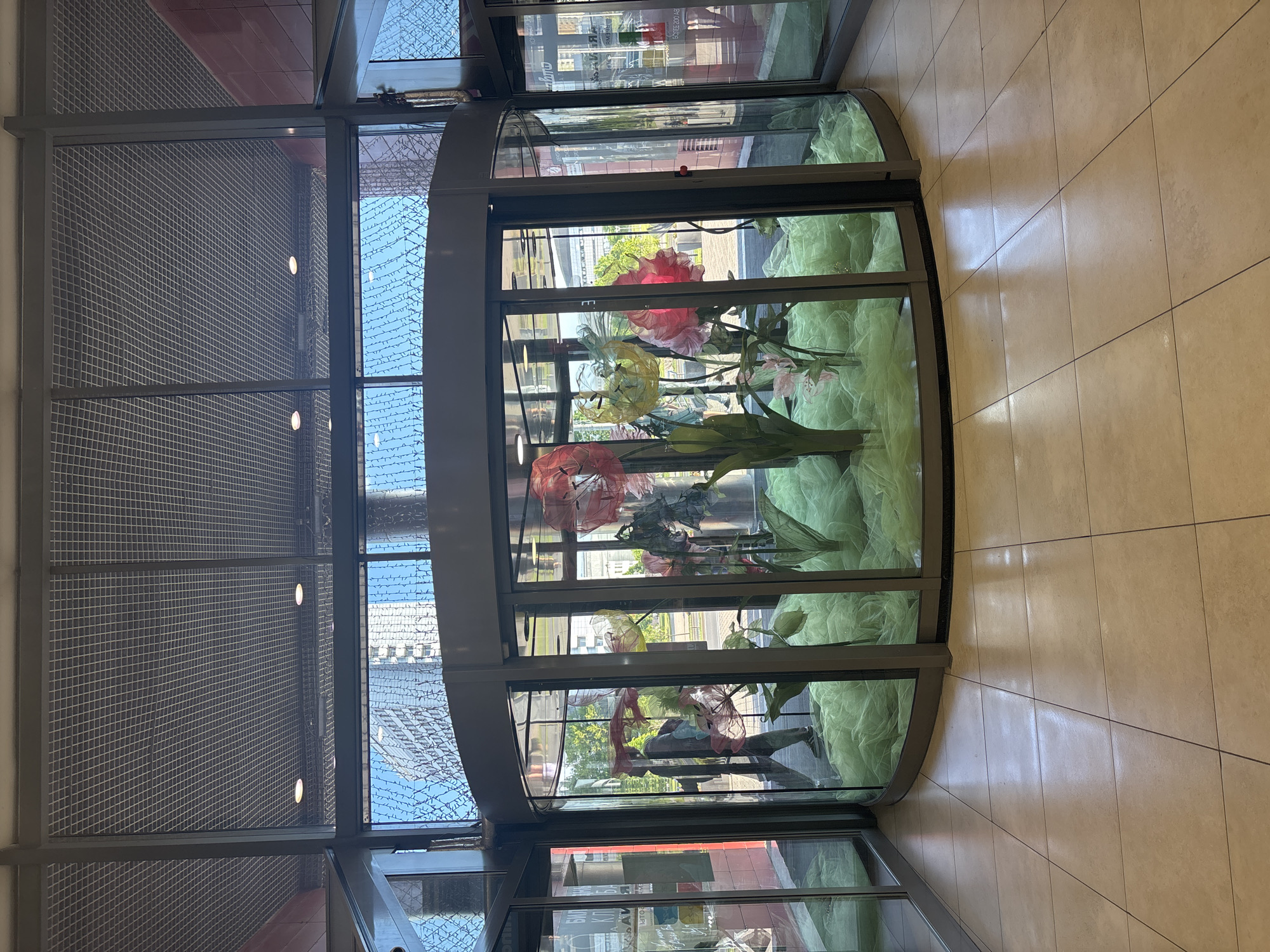 The image depicts the interior of a modern building, likely a commercial space, in Minsk, Belarus. A large, curved glass display case stands centrally, filled with an arrangement of artificial flowers and draped light green, translucent fabric, possibly tulle. The flowers are varied in shape and color, including prominent red, yellow, and pink blossoms resembling poppies or lilies, as well as lighter blue and white floral elements.
Through the clear glass of the display, an outdoor urban scene is visible, showing parts of other buildings, green trees, and what appears to be a street or pathway under a bright, clear sky. Reflections on the glass superimpose elements of the interior and distorted outdoor views. To the left of the display, a tall, grey metallic mesh or grating structure covers a wall, featuring several small, circular lights embedded within it. The floor to the right of the display is made of light-colored, square tiles, reflecting ambient light.
The scene is well-lit, indicating daytime. There are no clearly discernible people present in the image, although reflections might hint at distant figures. Reflected text on the glass is partially visible and mirrored; one segment appears to be "BOTTEGA," while other parts are indistinct, possibly including Cyrillic characters. The overall impression is one of a bright, contemporary setting with a decorative floral display.