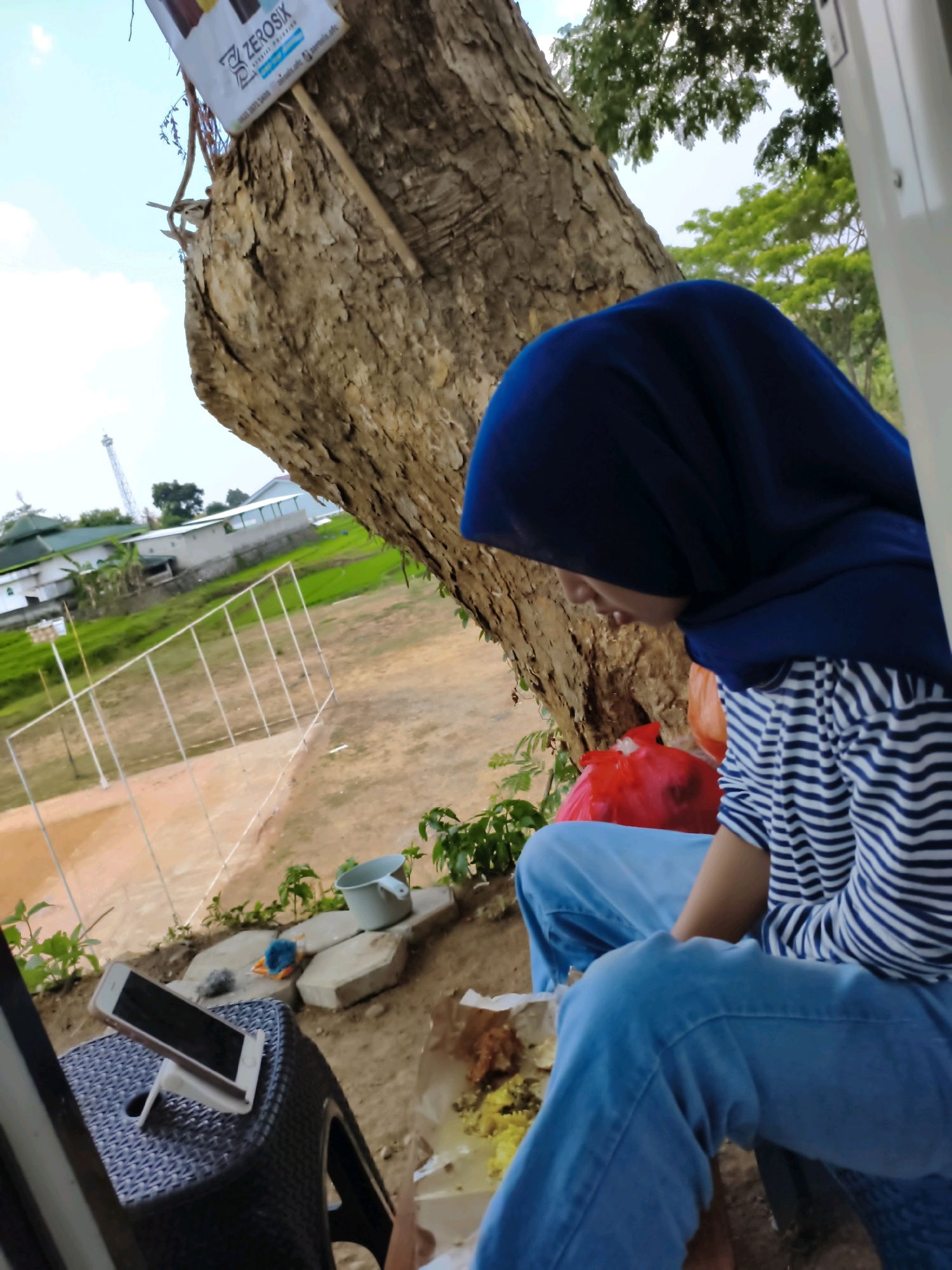 A person wearing a blue hijab and a striped shirt sits on a dark plastic chair, looking down at their food. They are wearing light blue jeans. In front of them on the chair is a smartphone in a white stand, displaying a black screen. To the left of the person is a large tree trunk with rough bark. A red plastic bag is visible behind the person. The ground is a mix of dirt and scattered stones, with some greenery growing around them. In the background, there is a field with a white fence, some buildings, and a distant tower. The overall scene appears to be outdoors, possibly in a rural or semi-rural area of Puhpelem, Indonesia.