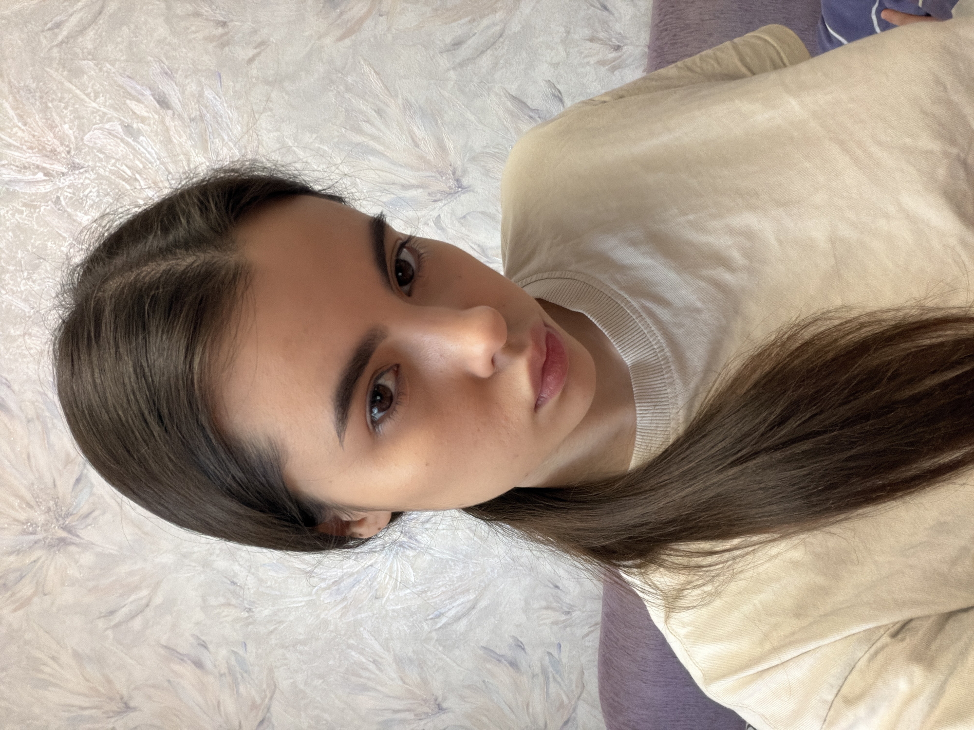 The media file presents a portrait of a young woman with long, dark brown hair and dark eyes. The original image is oriented on its side, depicting the woman with her head tilted to the right, looking directly towards the viewer with a neutral to slightly contemplative expression and slightly parted lips.
She has long, straight dark brown hair, parted approximately in the middle, flowing over her shoulders. Her eyebrows are well-defined, and her complexion appears clear. She is dressed in a light-colored, possibly beige or cream, long-sleeved top or sweatshirt with a ribbed crew neck, indicating casual attire.
The background features a light-toned wall with a distinct, textured pattern resembling stylized, wispy leaves or abstract brushstrokes in shades of white, light grey, and subtle purple. A small portion of a darker purple fabric object, possibly a cushion or blanket, is visible in the upper right part of the background. The lighting is soft and even, suggesting an indoor setting without strong directional light. No text is visible in the image. The accompanying crops provide closer detail of her hair, facial features, clothing, and the textured wall.