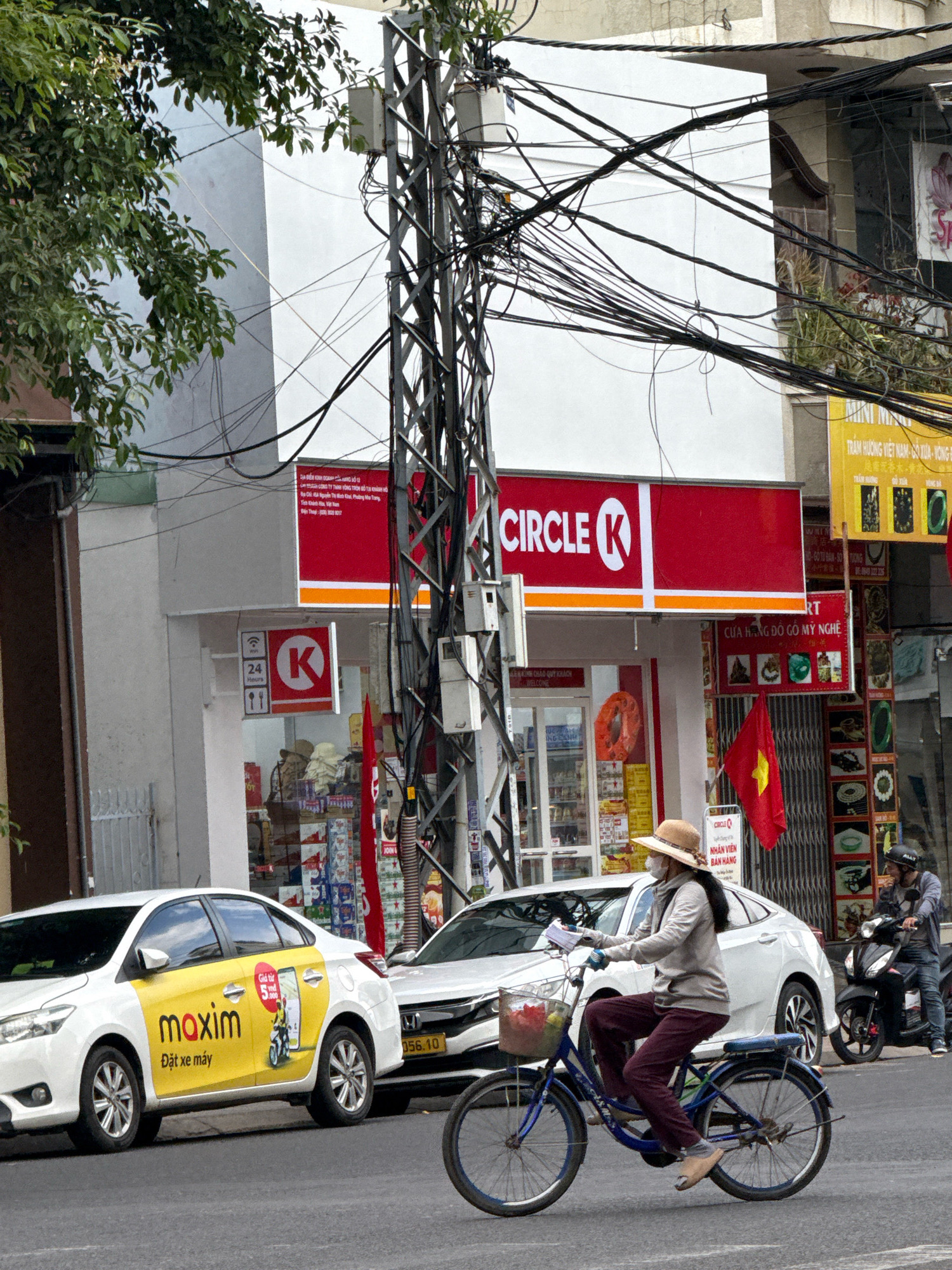 In an outdoor urban setting in Nha Trang, Vietnam, a white sedan with a yellow "Maxim" advertisement and Vietnamese text is parked on the left. To its right, a white SUV is also parked. In the center of the image, a woman wearing a tan straw hat, a grey hoodie, and maroon pants rides a blue bicycle with a basket containing a red bag. She is wearing a face mask and gloves. Behind the bicycle, a man on a black motorcycle with a rider wearing a grey jacket and helmet is visible, heading away from the camera. A red Circle K convenience store sign with white lettering is prominent on a building in the background, along with various other signs and wires. Red and yellow Vietnamese flags are also displayed. The street appears to be paved, and trees with green foliage are visible on the left.