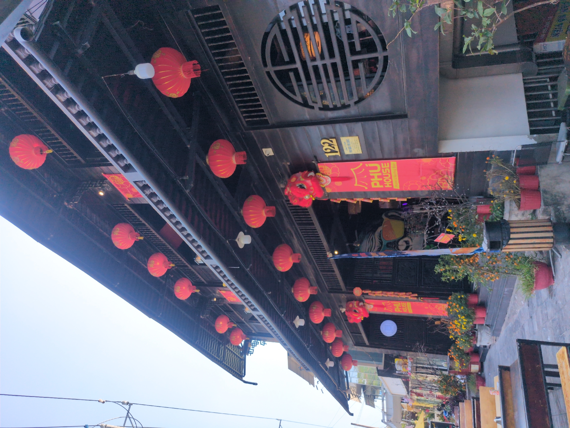 The image depicts a street scene in Phú Quốc, Vietnam, characterized by traditional Vietnamese architecture and decorations. Red lanterns are strung along the street, hanging from the eaves of the buildings. A prominent sign reading "PHÚ HOUSE" is visible, along with the number "122." Potted plants line the street, adding a touch of greenery. The overall impression is that of a festive and vibrant atmosphere, likely associated with a celebration or holiday.