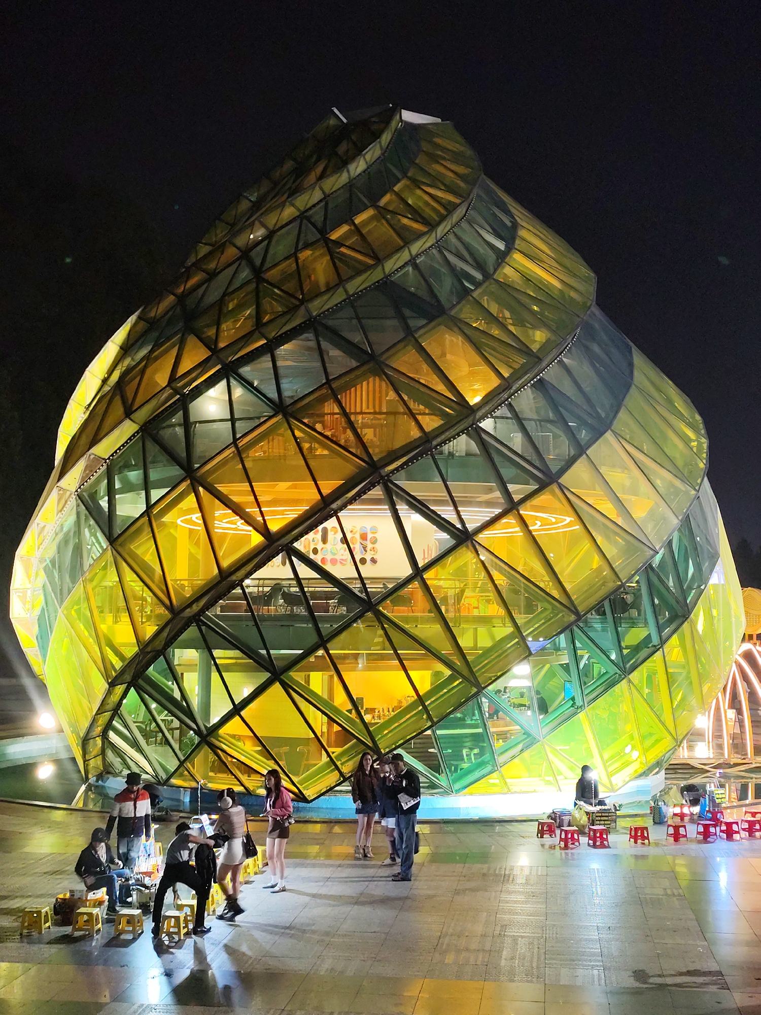 The image captures a lively night scene in Da Lat, Vietnam, centered around a striking, irregularly shaped glass building. This modern structure features a unique, spiraling design with a facade composed of numerous triangular and quadrilateral glass panels. These panels glow vibrantly, transitioning between shades of yellow and green, illuminated from within by warm artificial light that highlights internal spaces, including what appears to be a cafe or retail area with distinct circular neon light patterns.
In the foreground, on a paved outdoor plaza, several groups of people are gathered. On the left, an informal street vendor operates a stall, serving customers who are seated on small, yellow plastic stools. Another similar vendor with red plastic stools is visible on the right. People are seen standing, conversing, and engaging with the vendors, contributing to a bustling, casual evening atmosphere. The ground surface, possibly wet or reflective, mirrors some of the building's bright illumination. The dark night sky provides a stark contrast to the brightly lit building and ground activities. The scene depicts a blend of modern architecture and local street food culture at night. No prominent text is visible.