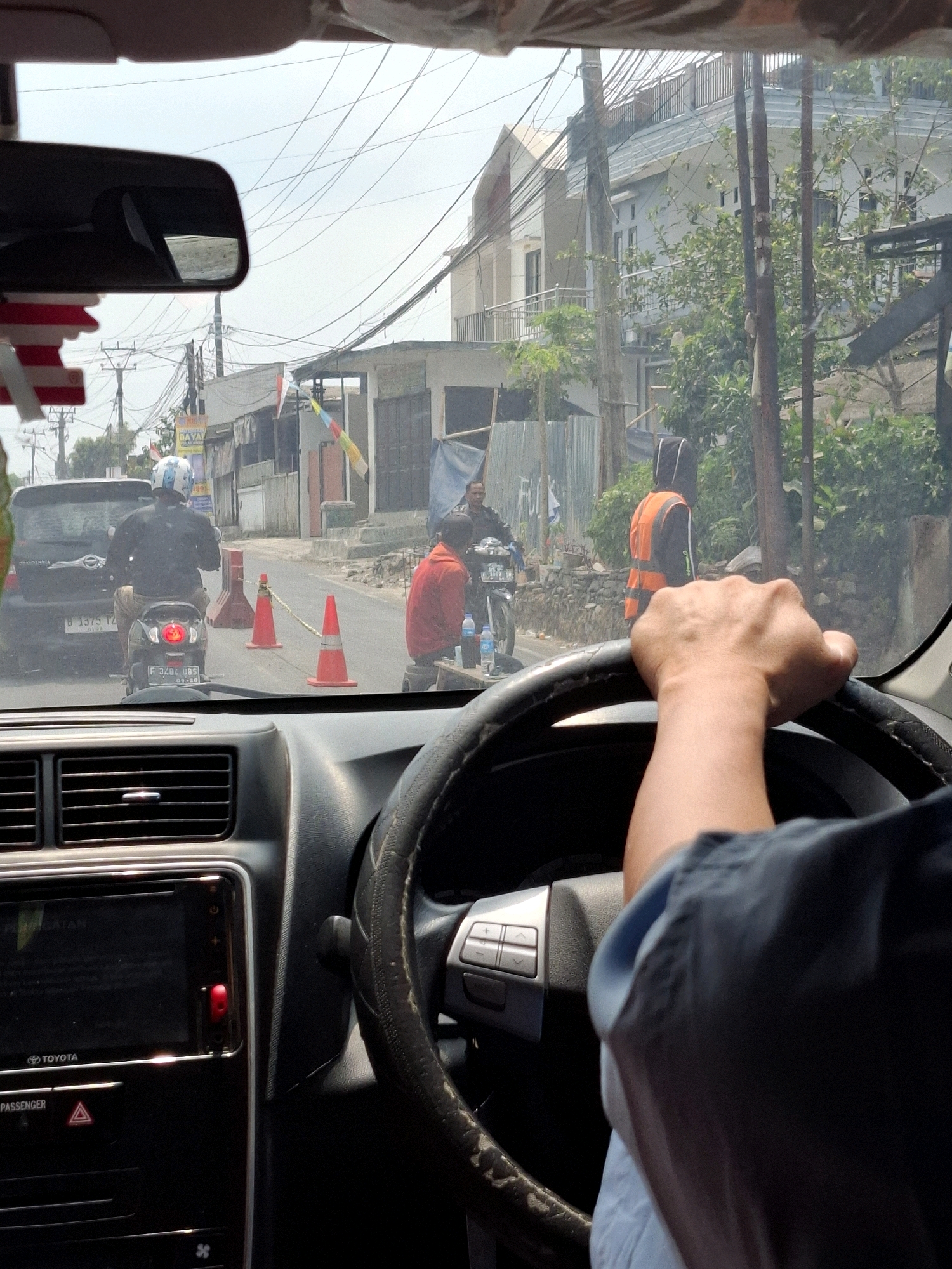 The image is taken from the perspective of a driver inside a car in Sukabumi, Indonesia. The driver's hands are visible on the steering wheel. Outside the car, traffic is moving on a street lined with buildings and trees. A motorcycle with a rider wearing a helmet is in front of the car, and a person wearing an orange vest is standing by the roadside. Orange traffic cones are placed on the street. The weather appears to be clear and sunny.