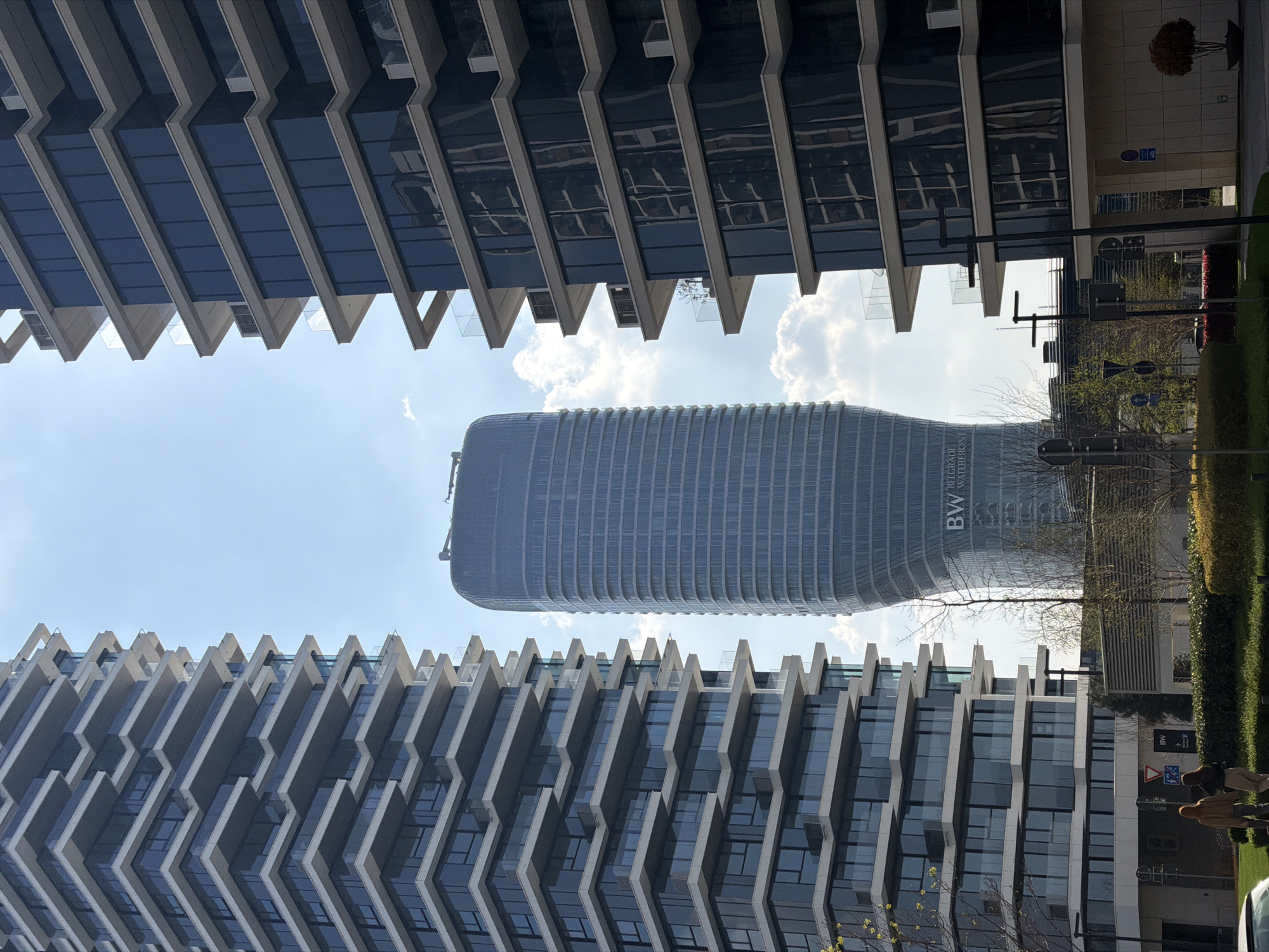 The image captures a view in Belgrade, Serbia, showcasing the Belgrade Waterfront complex. The frame is dominated by two modern buildings with a distinctive zigzag pattern on their facades, positioned in the foreground. Between these structures, the prominent BW (Belgrade Waterfront) building rises. The sky is visible above and around the BW building, suggesting a clear day with some clouds. On the right side of the image, there are trees, bushes, street signs, and a portion of a sculpture. The perspective suggests the photo was taken from a ground-level vantage point, looking upwards towards the buildings.