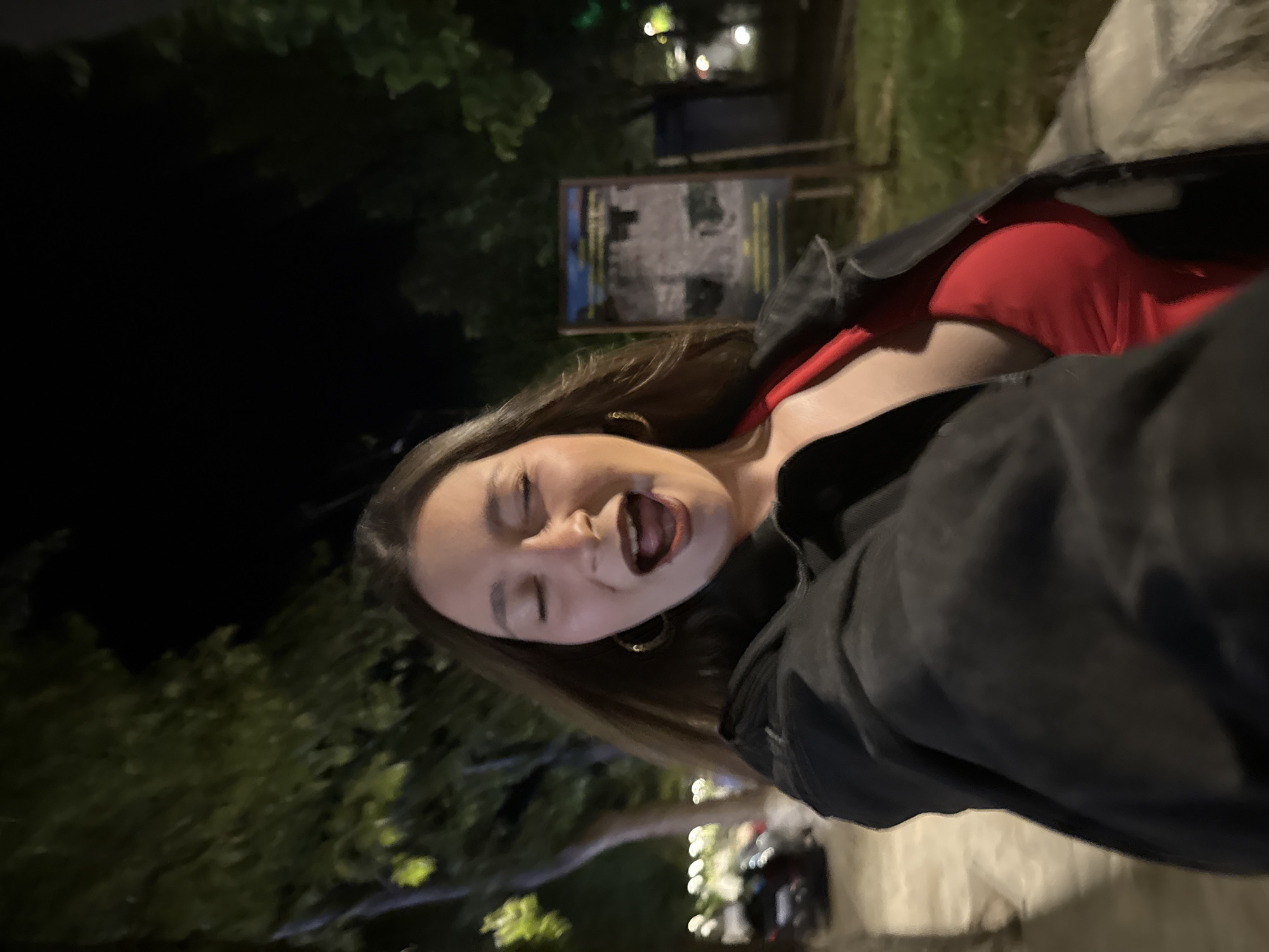 The image depicts a young adult female captured in a selfie at night in Sozopol, Bulgaria. She is positioned slightly off-center, with her head tilted to the right. Her eyes are closed or squinting tightly, and her mouth is wide open, revealing her teeth and dark lipstick. She has long brown hair, which falls over her shoulders, and wears gold-colored hoop earrings. Her attire includes a red garment, possibly a dress or top with a V-neckline, worn underneath a dark, likely black, denim-style jacket.
The background is an outdoor environment at night, dimly illuminated by artificial light sources creating significant bokeh effects. To the subject's left, out of focus, are trees with illuminated green foliage, a paved surface, and blurry white lights suggesting distant vehicles or streetlights. To her right, a rectangular informational sign mounted on a stand is visible, also out of focus, situated within a darker, vegetated area with more trees. A concrete curb or low wall runs across the lower right portion of the frame. The overall scene suggests a street or park area within a city.