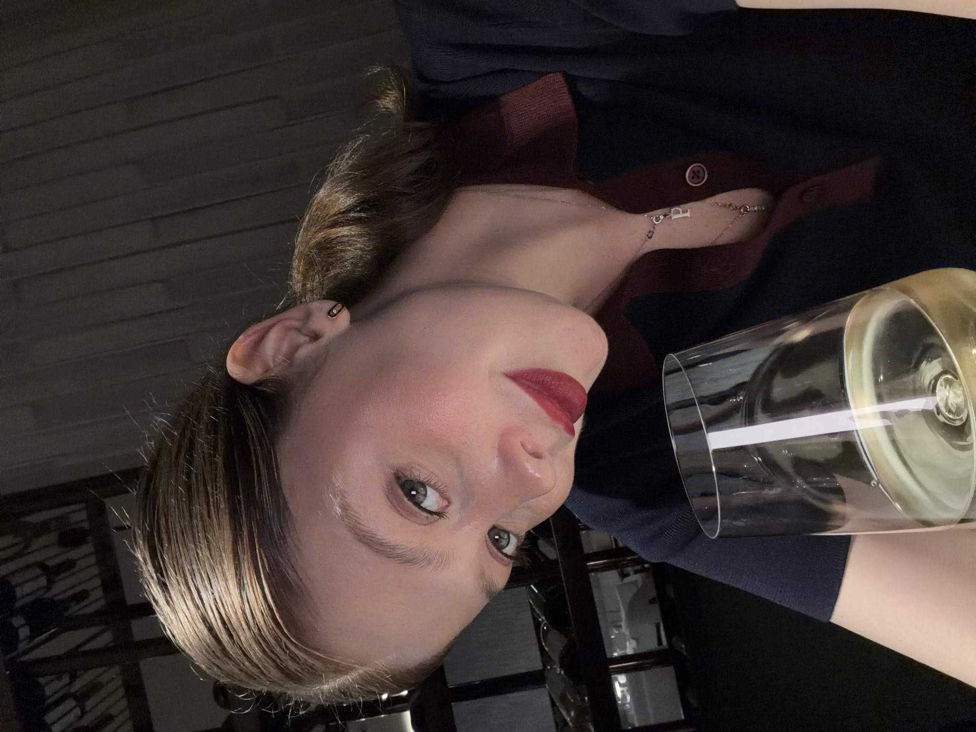 A woman is holding a glass of white wine. She is wearing a navy blue sweater with a burgundy collar. She has a silver necklace with a "P" pendant. She has red lipstick on. It is nighttime. The location is not identifiable as Tbilisi, Georgia.