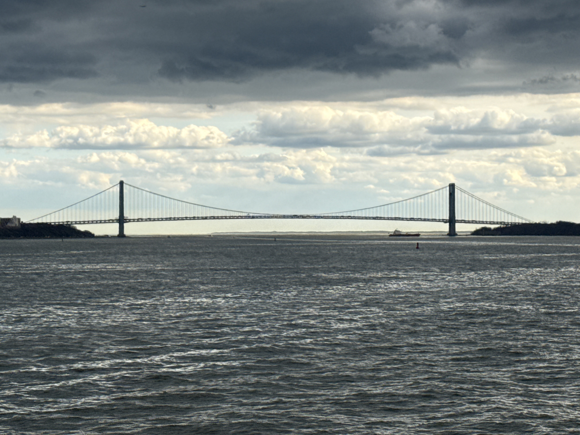 The image shows the Verrazano-Narrows Bridge spanning the Narrows strait in New York City. The view is likely from the vicinity of Bayonne, New Jersey. The scene features a large body of water with visible waves in the foreground. The bridge, a double-decked suspension bridge, stretches across the water connecting Staten Island and Brooklyn. The sky is overcast with dark, heavy clouds above lighter, more scattered clouds. There is a small vessel visible on the water near the bridge and a navigation buoy. Two landmasses frame the bridge on either side. The picture portrays a grey, slightly somber atmosphere.