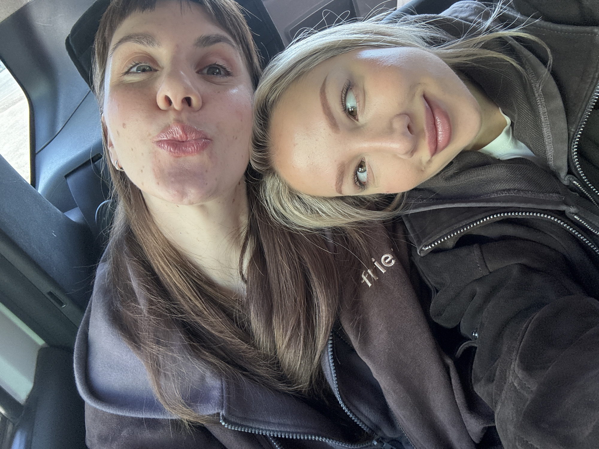 Two young women are posing close together for a selfie indoors, likely inside a vehicle. The woman on the left has brown hair, fair skin with some blemishes, and is making a puckered face. The woman on the right, leaning her head on the other woman, has blonde hair and is looking toward the left with a slight smile. Both are wearing dark-colored, zippered hoodies. The location is not evident in the image.
