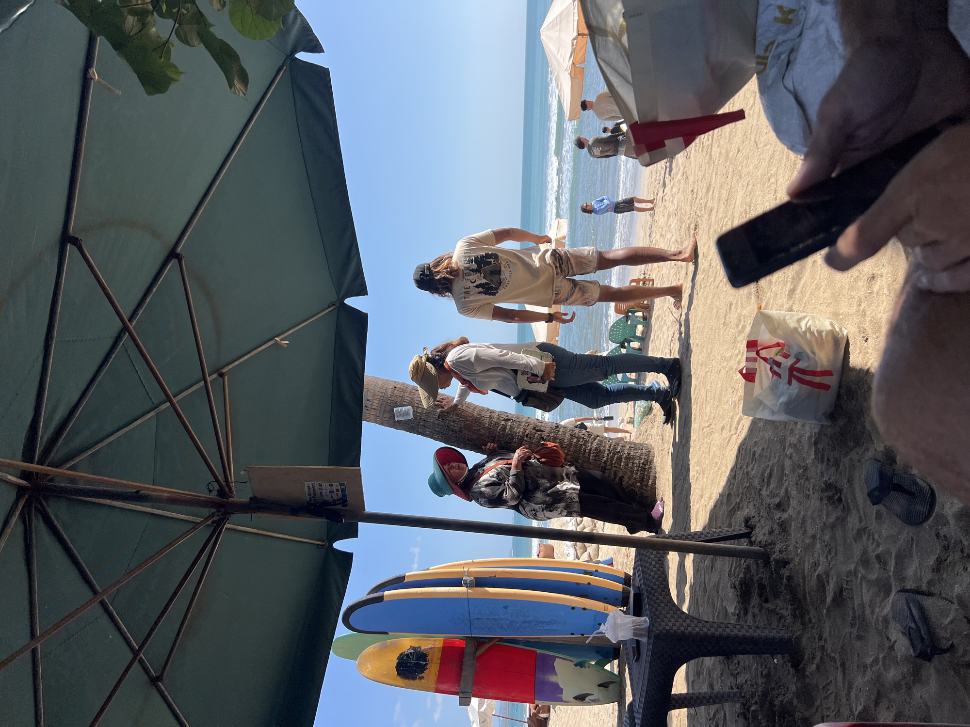 The image depicts a lively scene on a sandy beach in Kuta, Indonesia. In the foreground, a person's hands hold a smartphone, suggesting a moment of leisure or connection. Beside them, a white shopping bag with red ribbon is visible, indicating a purchase or a gift. The sandy ground is scattered with flip-flops and the shadow of an umbrella, hinting at a sunny day.
Further back, a dark-colored table holds a plastic cup with a straw, and several surfboards are propped up against a palm tree. Two women, one wearing a wide-brimmed hat and the other a visor, converse near the palm tree. The woman with the hat is dressed in a patterned shirt and dark pants, while the other wears a light-colored shirt and jeans.
Beyond them, on the beach, a man with long, wavy hair and a dark baseball cap stands facing the ocean. He wears a light-colored t-shirt and patterned shorts. Several other beachgoers are scattered around, some standing and some seated. Two orange beach umbrellas are set up further down the beach, and the gentle waves of the ocean create a serene backdrop. The sky is clear and blue, enhancing the tropical ambiance of the location.