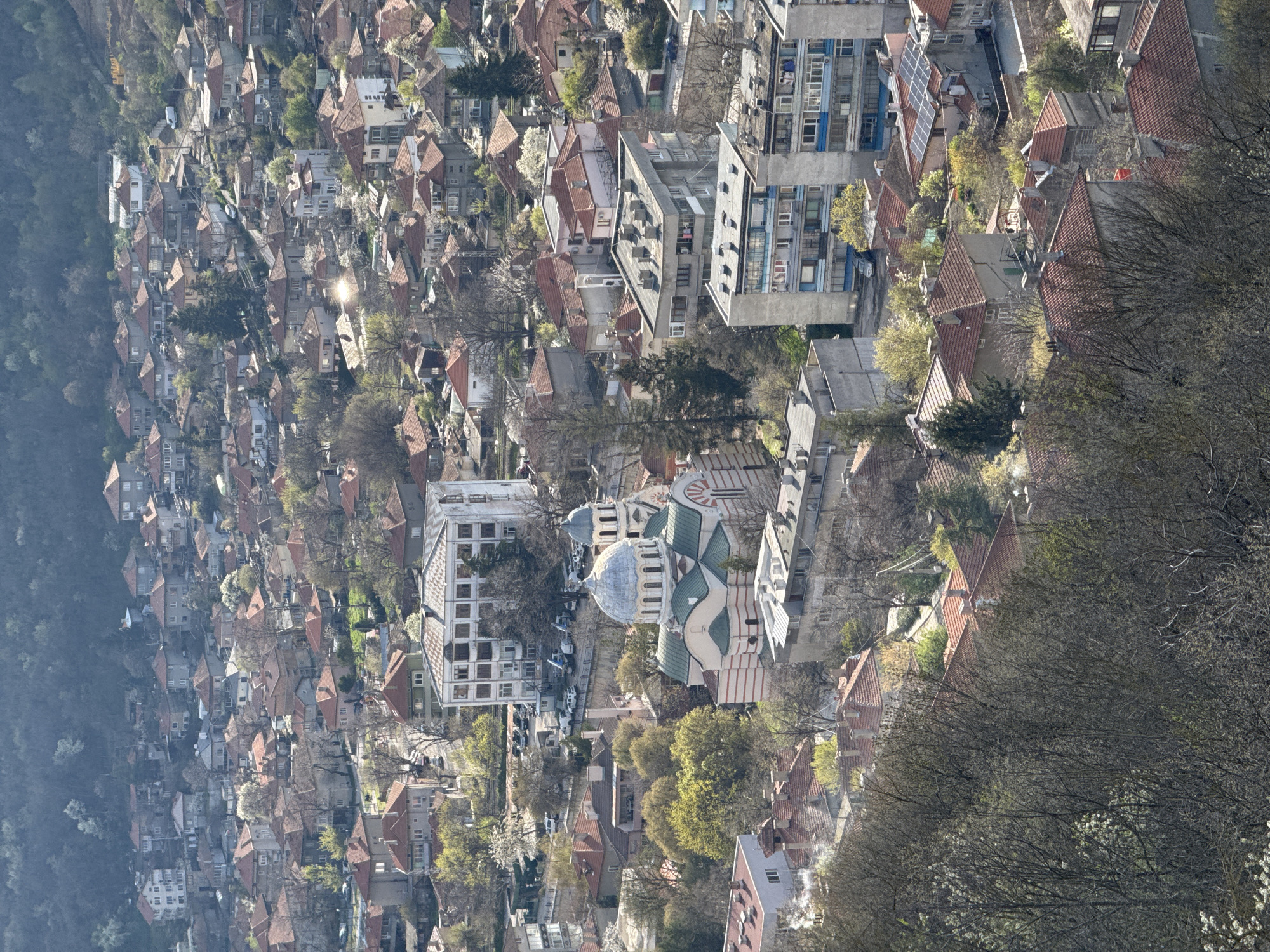 This aerial view shows a dense urban landscape in Provadia, Bulgaria. The scene is dominated by buildings of various sizes and styles, ranging from houses with red-tiled roofs to larger apartment blocks. A prominent church with a green and white striped design and a blue dome sits amidst the buildings. Bare trees are scattered throughout the cityscape, indicating it is likely late winter or early spring. The dense vegetation on the periphery suggests the town's edge borders a wooded area. The buildings are densely packed, with narrow streets visible between them.