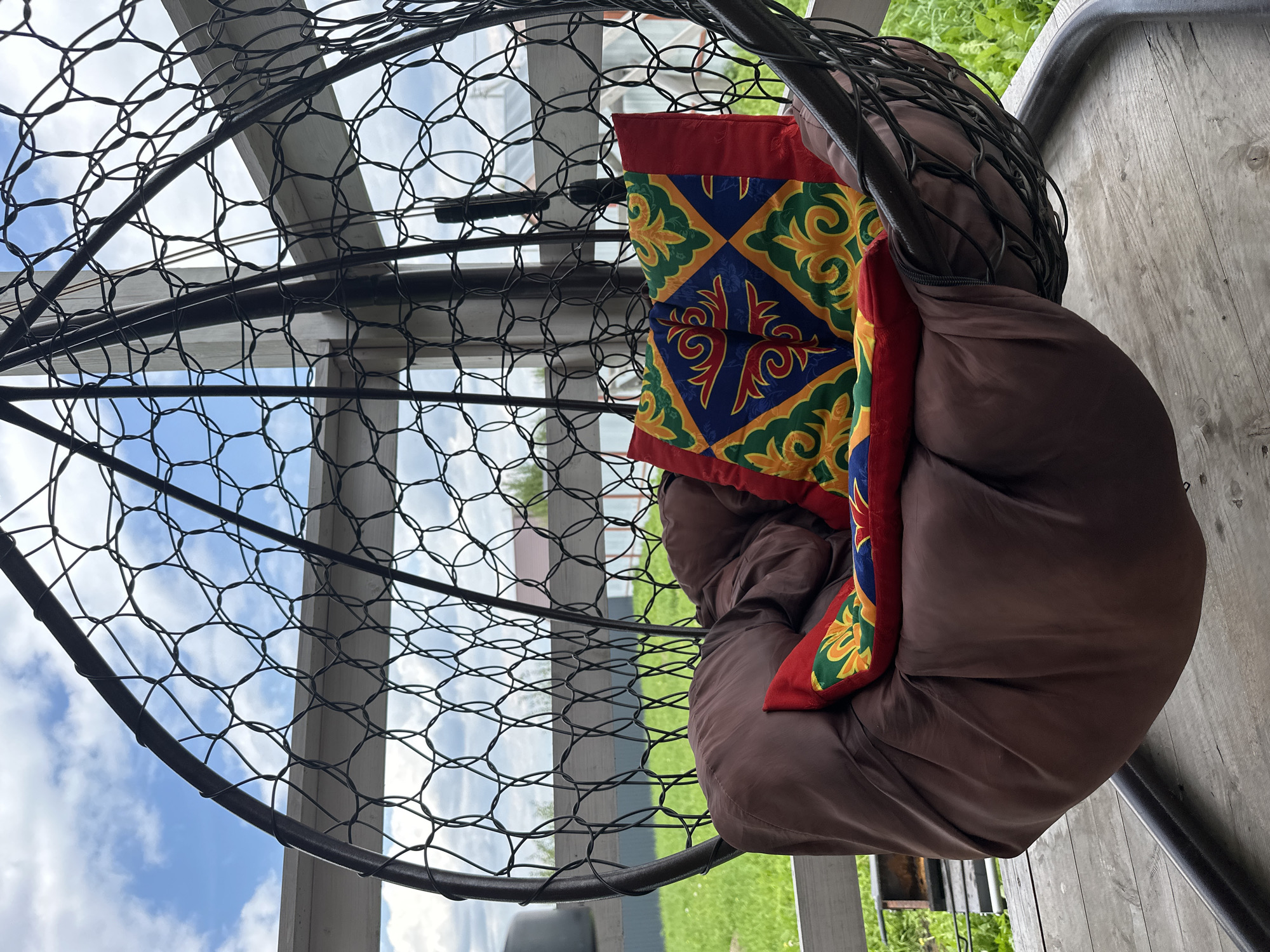 The image shows a hanging chair on a wooden deck with a background of green grass, a fence, and a cloudy sky. The chair is made of dark metal with a woven pattern and is filled with brown cushions and two colorful decorative pillows. The pillows have a geometric design with red, blue, yellow, and green colors. The scene appears to be in a residential outdoor area, possibly a backyard or patio. The location is specified as Taraskovo, Russia. There are no people visible in the image.