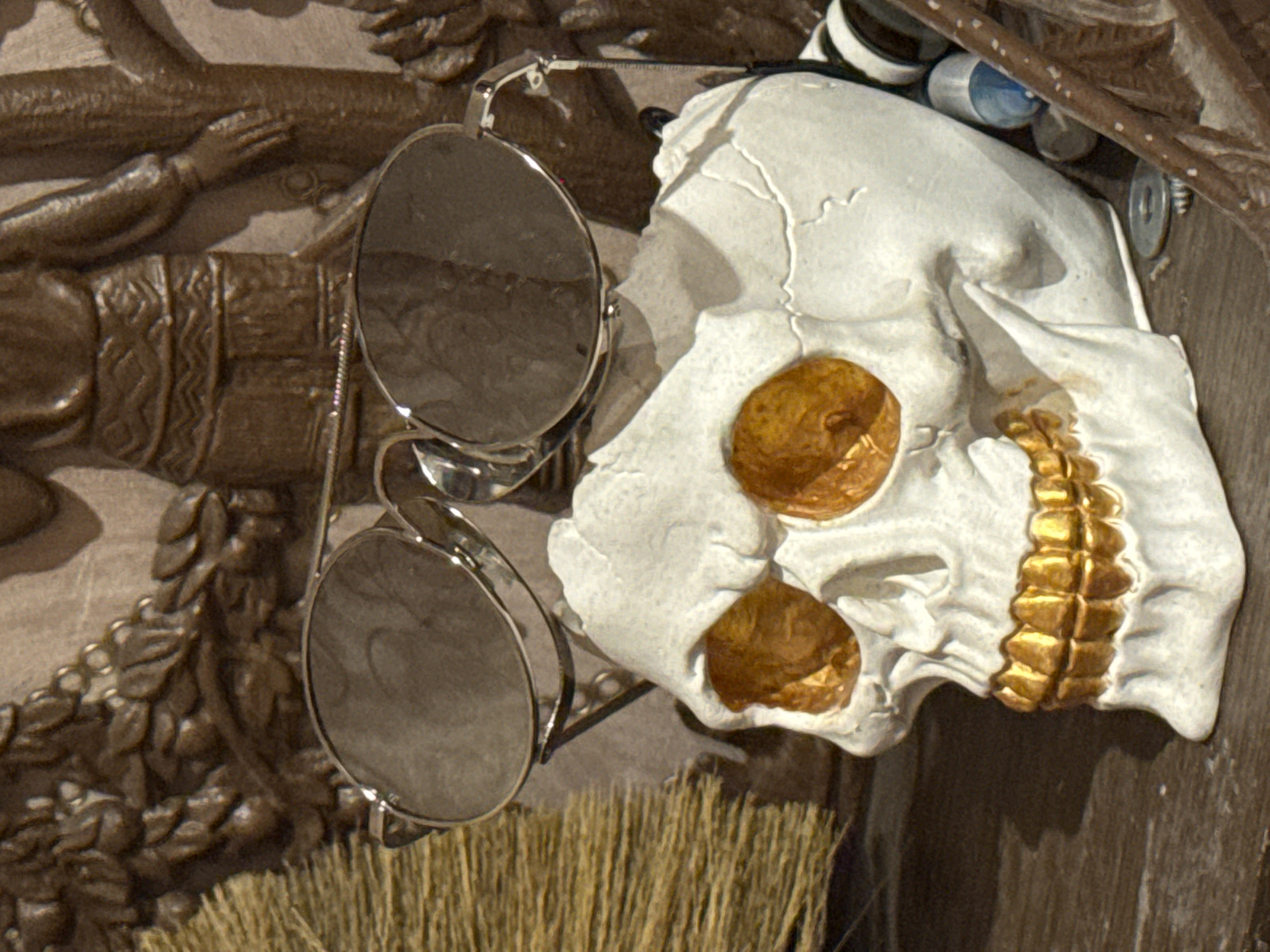 The image shows a close-up of a white skull adorned with gold accents in the eye sockets and along the jawline. Silver-rimmed sunglasses hang from the skull. The skull is positioned against a dark wooden background with carved figures of people. A bundle of straw is visible below the skull and glasses. The image was taken in Kladovitsy, Russia.