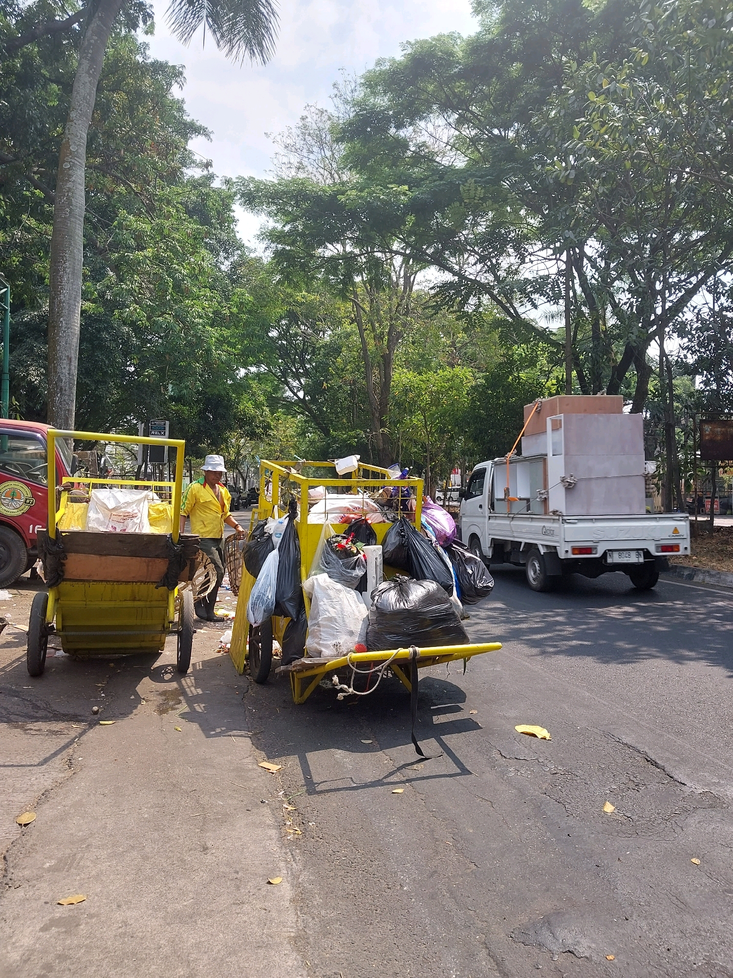In the city of Malang, Indonesia, on a sunny day, a man in a yellow shirt and white hat stands beside a yellow cart filled with trash bags. The cart has two wheels in the front and two wheels in the back. Next to it, another yellow cart is partially visible, also laden with trash bags and other debris. In the background, a white utility truck is parked on the side of the road, with a large, light-colored object secured on its bed. The scene is set against a backdrop of lush green trees and a clear sky, suggesting an outdoor environment, possibly a street or park area within the city. The road surface is asphalt, with some visible wear and tear.