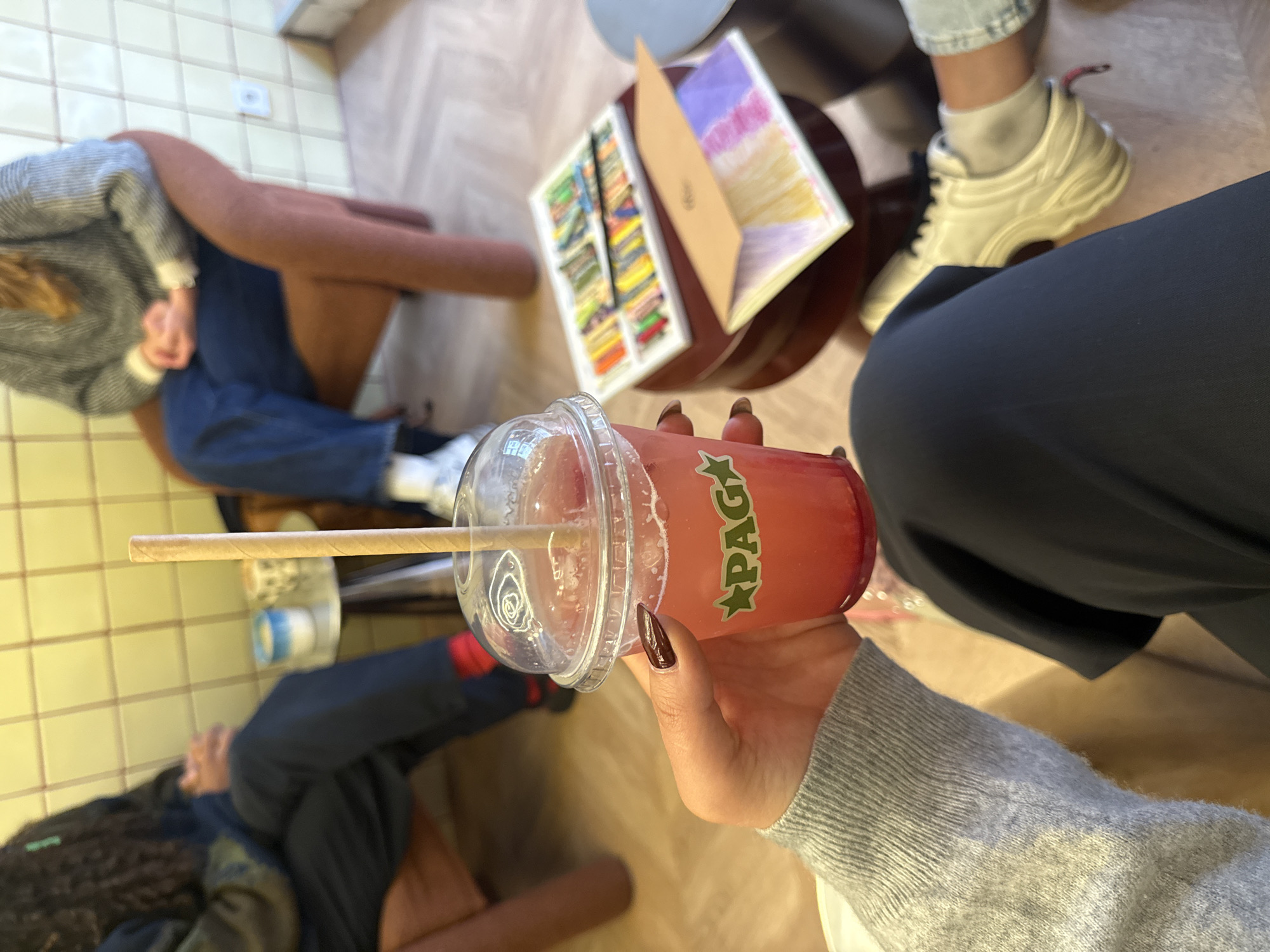A low-angle shot shows a hand holding a plastic cup containing a red drink with the word "PAGA" in green lettering, with a straw inserted. The hand has dark red nail polish. The forearm is clad in a gray sweater. The image also contains other people: a person wearing blue jeans and white shoes, a person wearing white sneakers with gray socks, and a person wearing black pants. A table holds a box of art supplies and drawing books. The image suggests a cafe scene with tiled floors. Based on the "PAGA" brand on the drink, the location may be in Paris, France, where this cafe chain has locations.