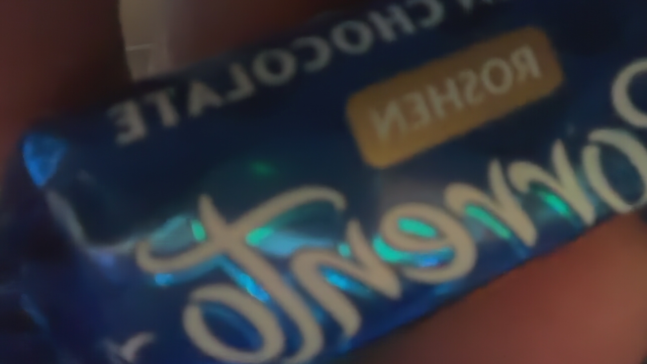 A person's hand is holding a blue chocolate bar wrapper in an indoor setting. The image is blurry and out of focus, making some details difficult to discern. The wrapper has a glossy or holographic finish, reflecting iridescent blue and green light, particularly around the large, stylized text.
Visible text on the wrapper, primarily inverted, includes:
* "CHOCOLATE" is clearly visible across the top, written in white uppercase letters.
* A golden rectangular box contains the Cyrillic word "РОШЕН" (ROSHE(N)), indicating the Roshen brand.
* Below "CHOCOLATE," a large, stylized script text, possibly "Jonny Krock," is featured prominently in white with holographic reflections.
* Other partial, inverted letters are visible on the left side of the wrapper, but they are too blurry to be definitively identified beyond fragments like "LATE" which could be part of a larger descriptor.
The activity taking place is simply someone holding the chocolate bar. The setting is indistinct, with a dark, out-of-focus background suggesting an interior environment. No specific time of day, weather, or location cues are visible within the image itself. The overall impression is a casual, close-up shot of a chocolate product.