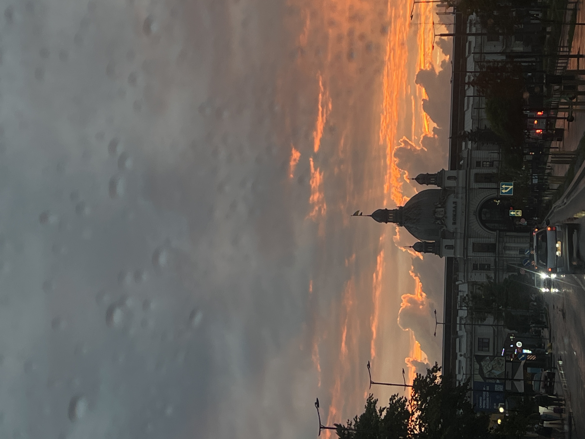 The image depicts a sunset over the Lviv Railway Station (Vokzal Lviv) in Lviv, Ukraine. The sky is filled with dramatic clouds tinged with vibrant orange and yellow hues, indicative of a sunset. The station building, with its distinctive domes and architectural details, is visible in the mid-ground. In the foreground, a wet street suggests recent rain, with cars driving and streetlights illuminating the scene. Several trees line the street, and some pedestrians are visible on the sidewalks. The overall atmosphere is one of a city transitioning from day to night under a striking sky.