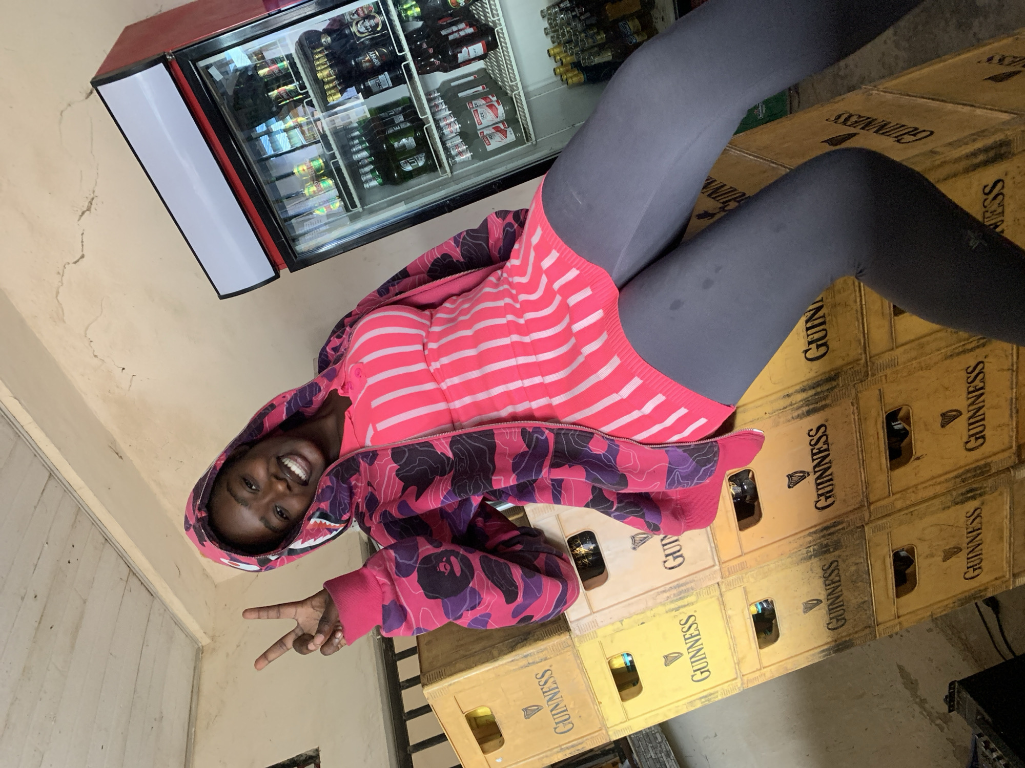 A young Black woman is pictured in an outdoor setting, leaning against a stack of yellow Guinness crates. She is wearing a pink and purple camouflage hoodie over a pink and white striped dress, and grey leggings. She is smiling and holding up two fingers in a peace sign. To her right is a red and white commercial refrigerator filled with bottles of beverages. The background shows a light-colored wall and a wooden ceiling. The scene appears to be a local business or bar in New Stadium, Nigeria.