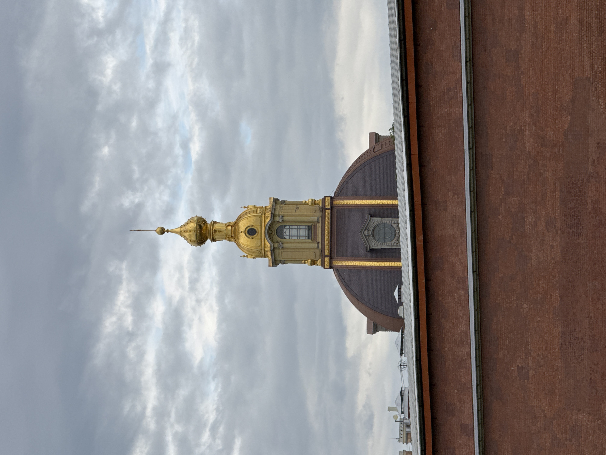 The image shows a golden dome of a building against a cloudy sky. The dome is ornate and has a spire on top. Below the dome, there is a dark, reddish-brown dome with some decorative elements and windows. In the foreground, there is a large brick wall. The view is from a medium distance. This is likely a view of a historical building in Saint Petersburg, Russia, possibly within a complex like the Peter and Paul Fortress, given the architectural style.