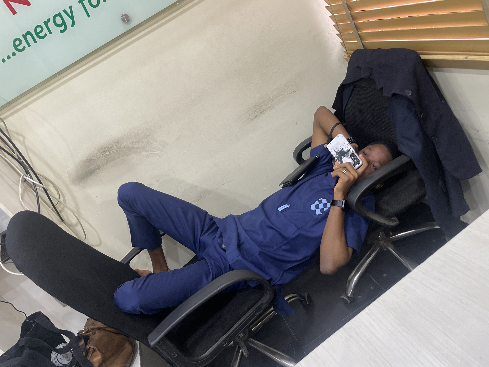 A young man in a dark blue uniform is reclined in a black office chair, with his legs over the backrest. He is holding a white mobile phone with a black design on it, and appears to be looking at it. A dark navy jacket is draped over the chair's armrest and on the floor. The background consists of a light-colored wall with some dark smudges, a section of vertical wooden blinds, and office equipment including wires. A light-colored desk is partially visible in the lower left corner. The scene is likely an office environment in Ikeja, Nigeria.