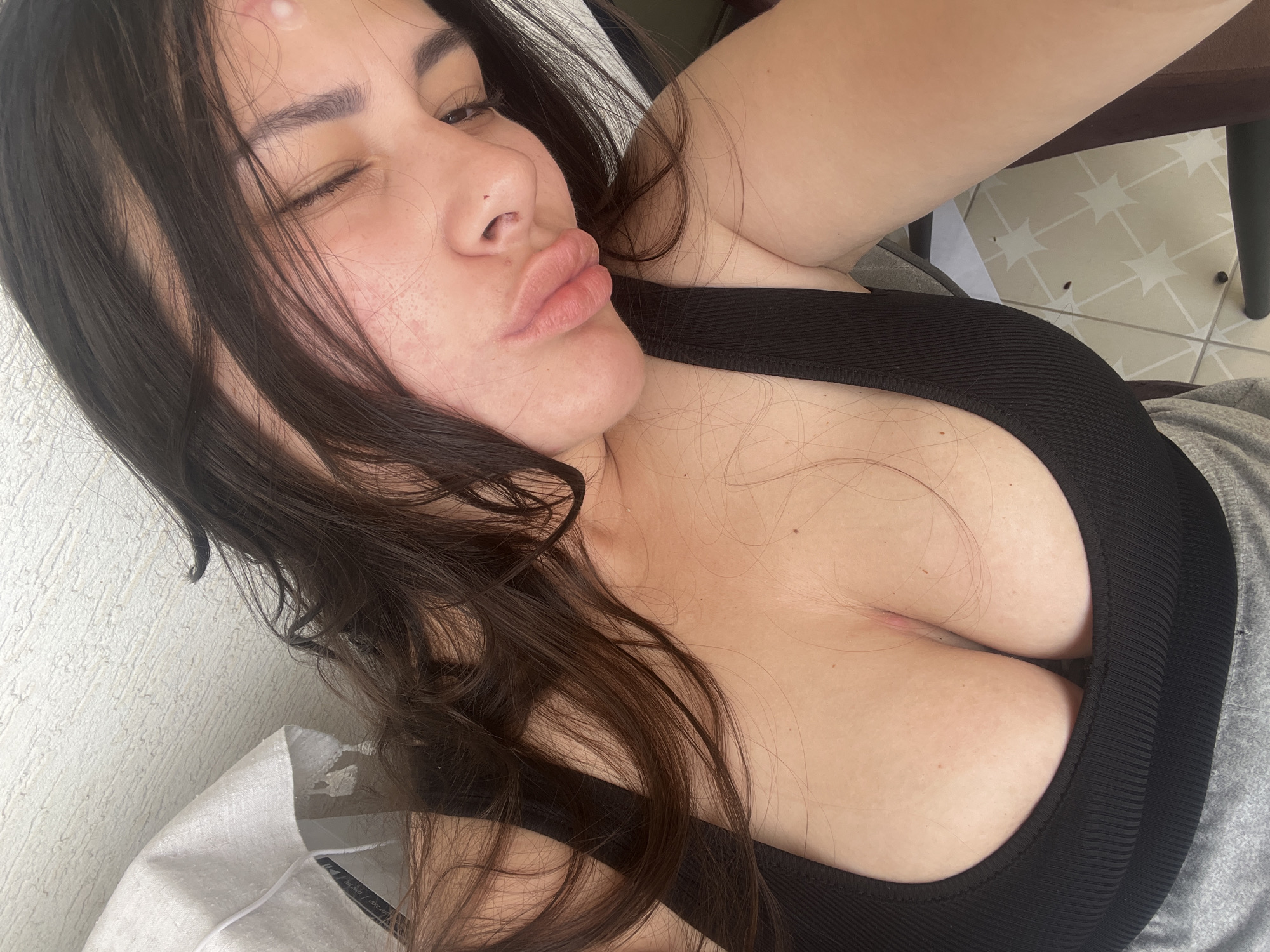 A woman with long, dark hair is lying down and looking at the camera with pursed lips and one eye partially closed. She has a blemish on her forehead and some redness on her cheek. She is wearing a black ribbed top that reveals a significant amount of cleavage. She is lying on a light-colored surface, possibly a bed or couch, with a gray garment nearby. The background includes a textured wall and a glimpse of a chair and tiled floor, suggesting an indoor environment. There is no information to suggest that the image was taken in Kiryat Bialik, Israel.
