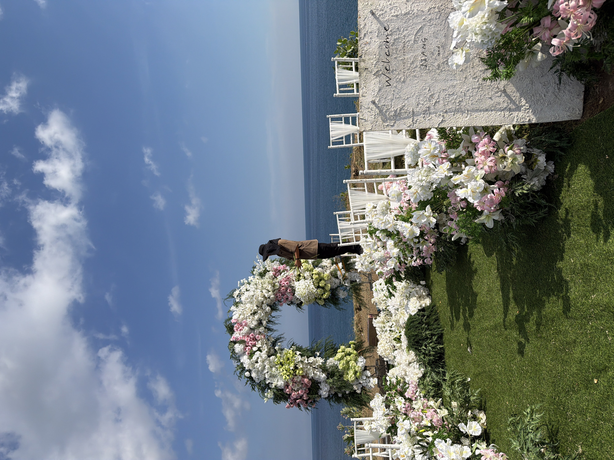 This image depicts an elegantly decorated outdoor event space, likely set up for a wedding ceremony, overlooking the ocean in Kuta Selatan, Indonesia. The scene is dominated by extensive floral arrangements in shades of white, pink, and light green, combined with lush green foliage.
A large, circular floral arch, resembling a wreath, is a central feature. Standing next to this arch is a person, dressed in a brown shirt and black trousers or skirt, with a black head covering. They appear to be making final adjustments to the floral decorations. To the right, several rows of white chairs, some adorned with draped white fabric, are arranged, facing the expansive blue ocean. These chairs are bordered by more dense floral displays that complement the arch.
Further to the right, a white, textured sign or wall is visible, bearing black scripted text that reads "Welcome JIANG and WANG." The ground beneath the event setup is covered with neat green artificial turf.
The background features a vast, calm blue ocean meeting a clear blue sky, which has scattered white cumulus clouds. The bright, even lighting indicates it is daytime, likely late morning or early afternoon, with sunny and fair weather. The overall impression is one of meticulous preparation for a special celebratory occasion in a picturesque, cliffside setting.