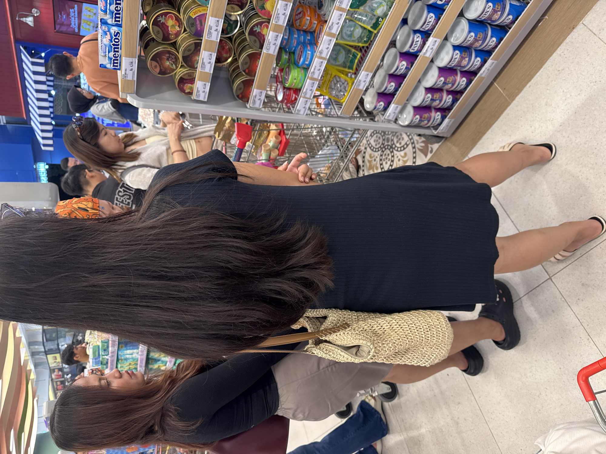 Here is a detailed description of the image:
The image captures a scene inside a store, likely a supermarket, possibly located in Kuta, Indonesia, although that location is not verifiable from the image. The perspective is from a high angle, looking down at the customers and products.
**People:**
- Two women are in the foreground. One has long, dark, straight hair and is wearing a black dress and black Crocs. She carries a woven bag with a brown strap. The second woman has medium-length brown hair and wears a black long-sleeved shirt and a beige skirt.
- In the background, several other shoppers are visible, some browsing the shelves.
**Objects:**
- Supermarket shelves are filled with various canned and packaged food items. Specific brands visible include "Mentos" and "Fox's" candies.
- A shopping cart is partially visible on the right.
**Environment:**
- The flooring is light-colored, possibly tiled.
- The store appears to be well-lit, with a variety of merchandise on display.
- Price tags are visible on the shelves, including one that says "Rp. 4,530," indicating Indonesian Rupiah currency.
**Actions/Interactions:**
- Shoppers are browsing and presumably selecting items from the shelves.
**Summary:**
The image depicts a busy scene inside a supermarket with shoppers examining the available products.