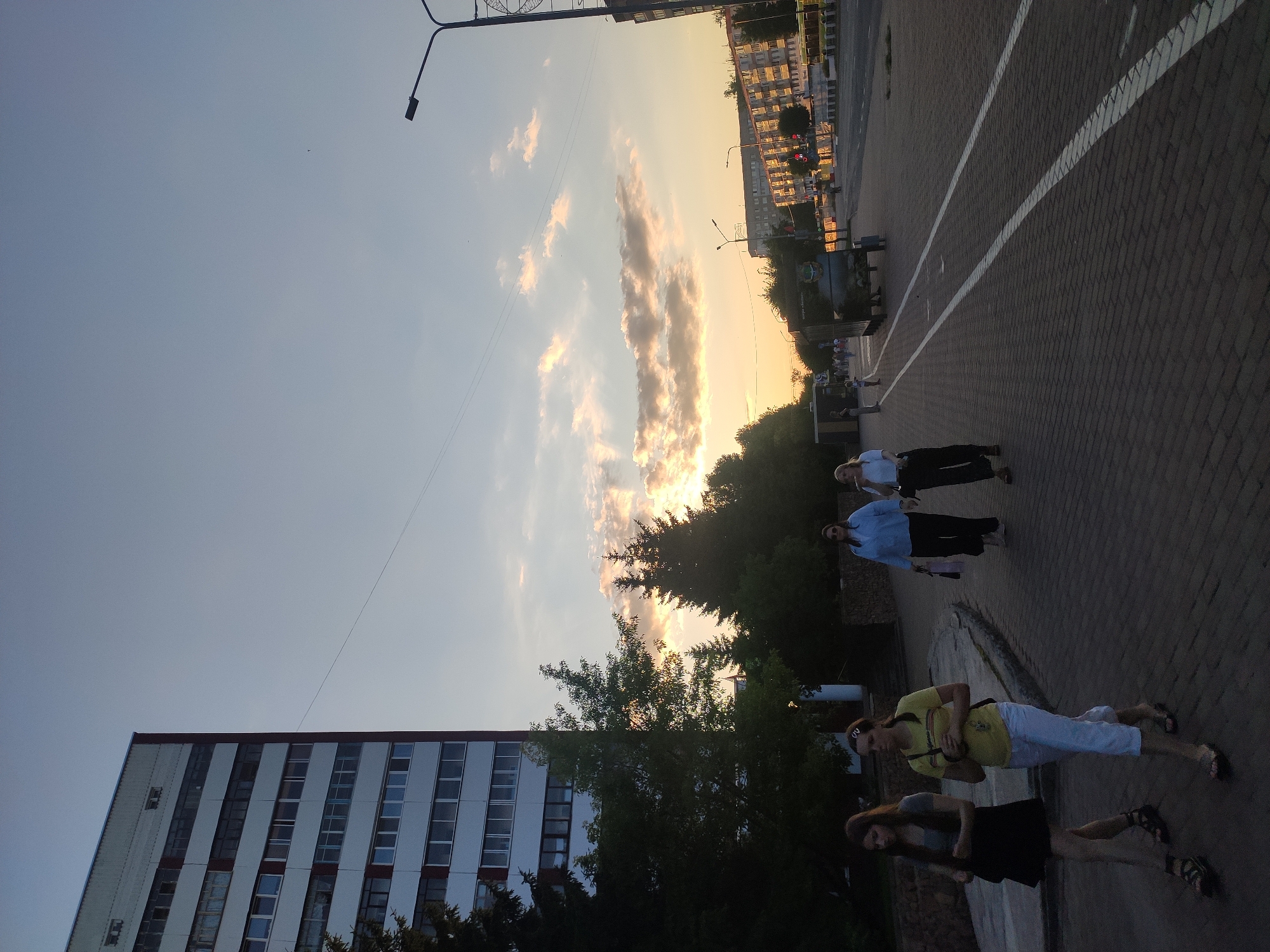 The image depicts a street scene in Navapolatsk, Belarus, during what appears to be late afternoon or early evening, with the sky displaying a blend of blue and warm sunset colors, punctuated by scattered clouds. On the left side of the frame, a tall, modern building with a white and red facade dominates the background, featuring numerous windows arranged in horizontal rows. Adjacent to the building and extending towards the right are trees, some appearing as evergreens and others as deciduous, silhouetted against the brighter sky.
In the foreground, a paved walkway or sidewalk, constructed from brick-like pavers, runs diagonally from the lower left to the mid-right of the image. Several individuals are visible walking on this pathway. Closer to the left foreground, two young women are walking side-by-side. The woman on the left wears a black skirt and a dark top, while the woman on the right is dressed in a yellow top and white pants. Further down the path, two more women are walking away from the camera. One is wearing a blue shirt and dark pants, and the other a white top and dark pants.
The right side of the image shows a road with passing vehicles, suggesting an urban environment. Streetlights are present along the road. Further in the distance, more buildings can be seen, some of which are multi-story residential structures. The overall impression is that of a typical urban street, possibly a public promenade or a pedestrian area alongside a main road, during a time of day when people are out and about. The lighting suggests the sun is low on the horizon, casting long shadows and bathing the scene in a warm glow.