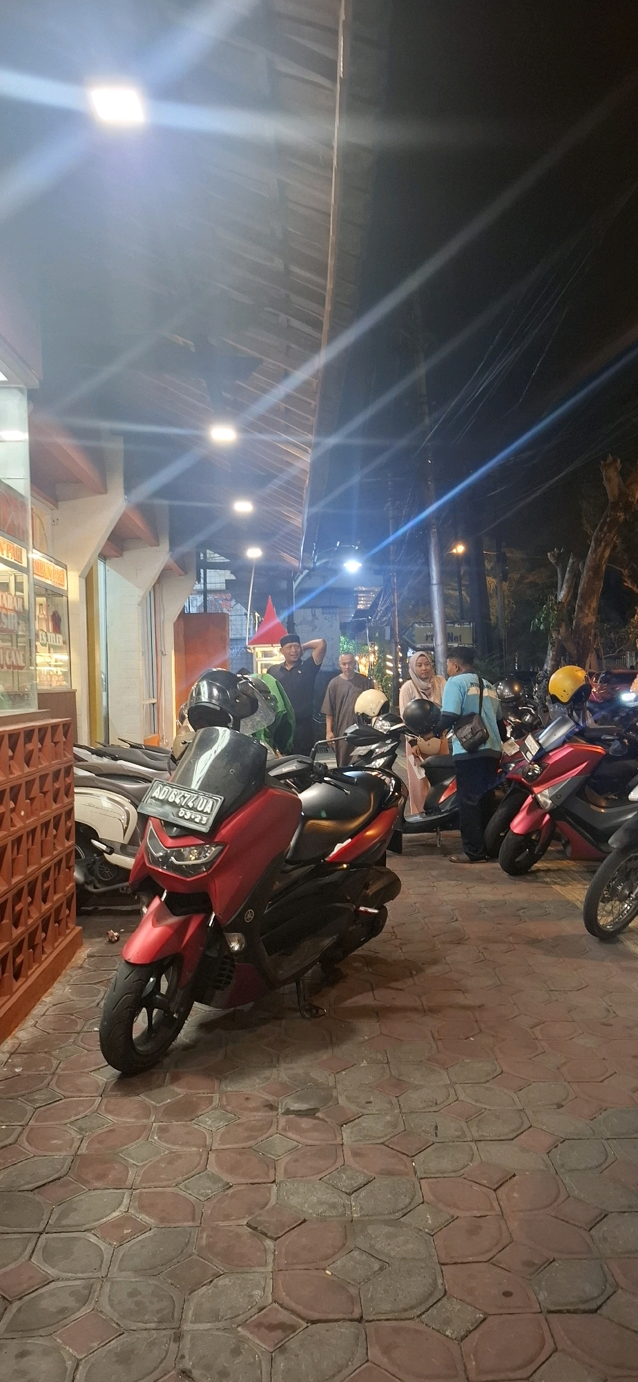The image depicts a street scene at night in Surakarta, Indonesia, likely in a commercial or entertainment area given the presence of food stalls and parked motorbikes. The foreground is dominated by a red Yamaha Nmax scooter with a black helmet on its seat. The scooter has a license plate reading "AD 8472 UJA". To the left of the scooter, a section of a food stall with a red brick patterned wall is visible.
In the mid-ground, several people are standing near other parked motorbikes. A man in a black uniform with a cap is touching his head, seemingly engaged in conversation with others. Another man in a grey t-shirt stands nearby, and a woman wearing a beige hijab and a light pink dress is talking to a man in a blue t-shirt. Several other motorbikes, including a pink and black scooter with a yellow helmet, are parked in the background.
The environment features a covered walkway with exposed wooden beams and overhead lights that cast strong beams. Power lines are visible against the dark sky. The ground is paved with interlocking hexagonal and diamond-shaped tiles in shades of red and gray, some of which appear wet. The overall impression is a lively street corner at night, bustling with activity.
