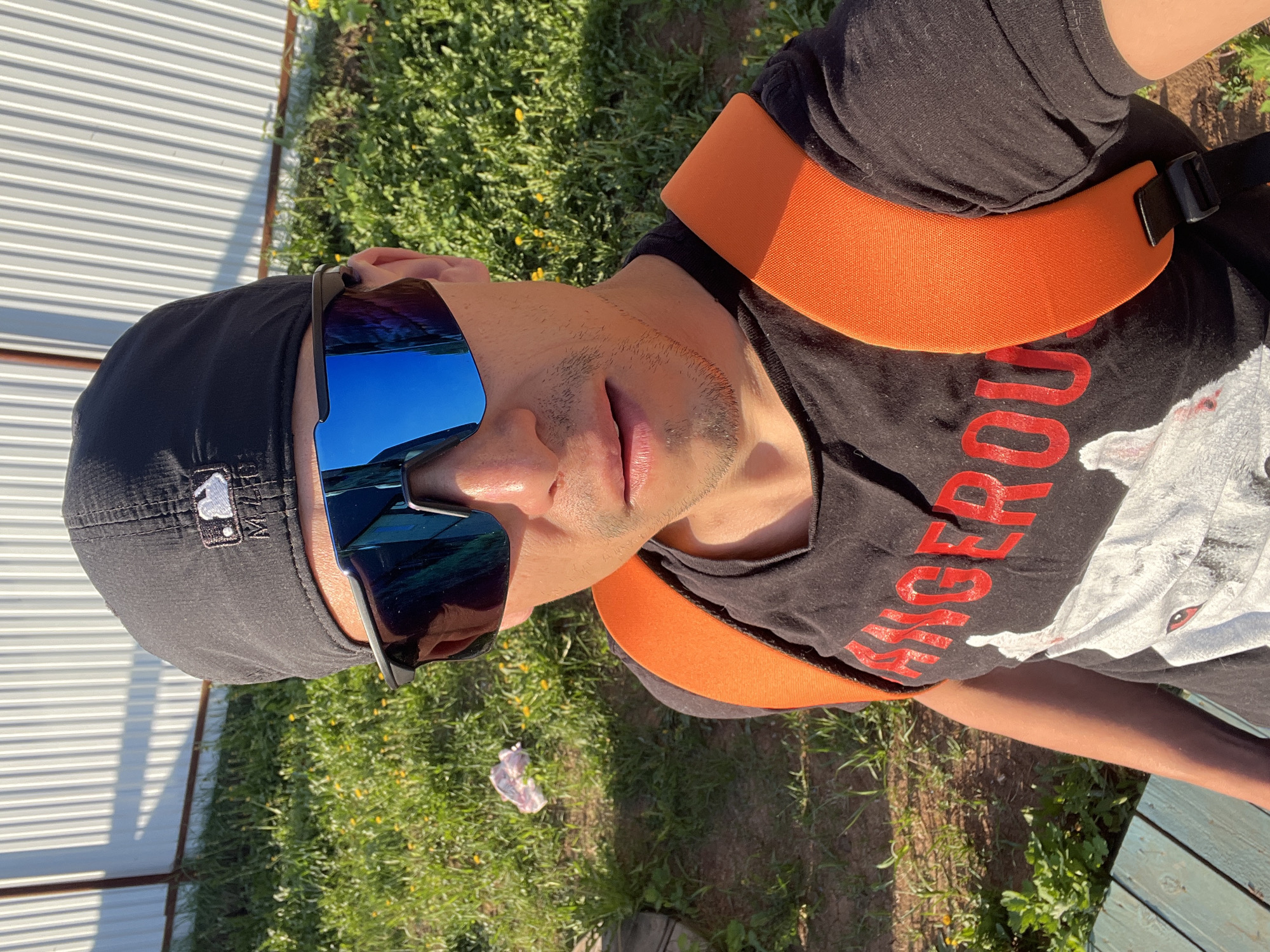 A man wearing a black t-shirt with red lettering, a black baseball cap with an MLB logo, and blue mirrored sunglasses is outdoors, possibly sitting on a wooden bench. He has an orange padded harness across his shoulders and chest. The background includes grass and a white metal fence. The location is suspected to be in the city of Chaikovsky, Russia, based on available data.