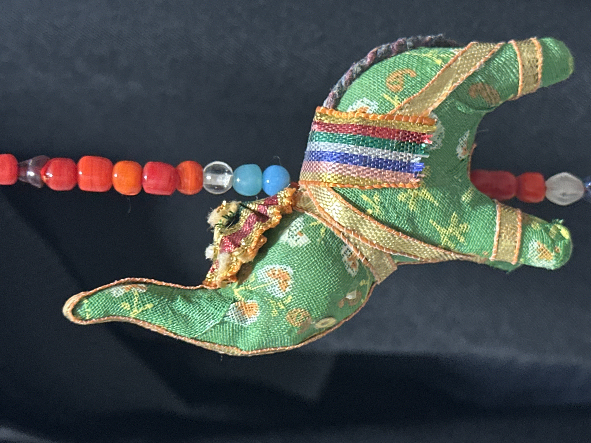 A close-up, indoor photograph shows a green fabric elephant ornament with gold trim and a colorful striped saddle, hanging against a dark, out-of-focus background. The elephant has a trunk raised upwards. A string of red, blue, and clear beads is visible hanging above and behind the elephant. The lighting appears to be artificial, creating some shadows on the elephant's surface. The photograph was taken in Nevinnomyssk, Russia.