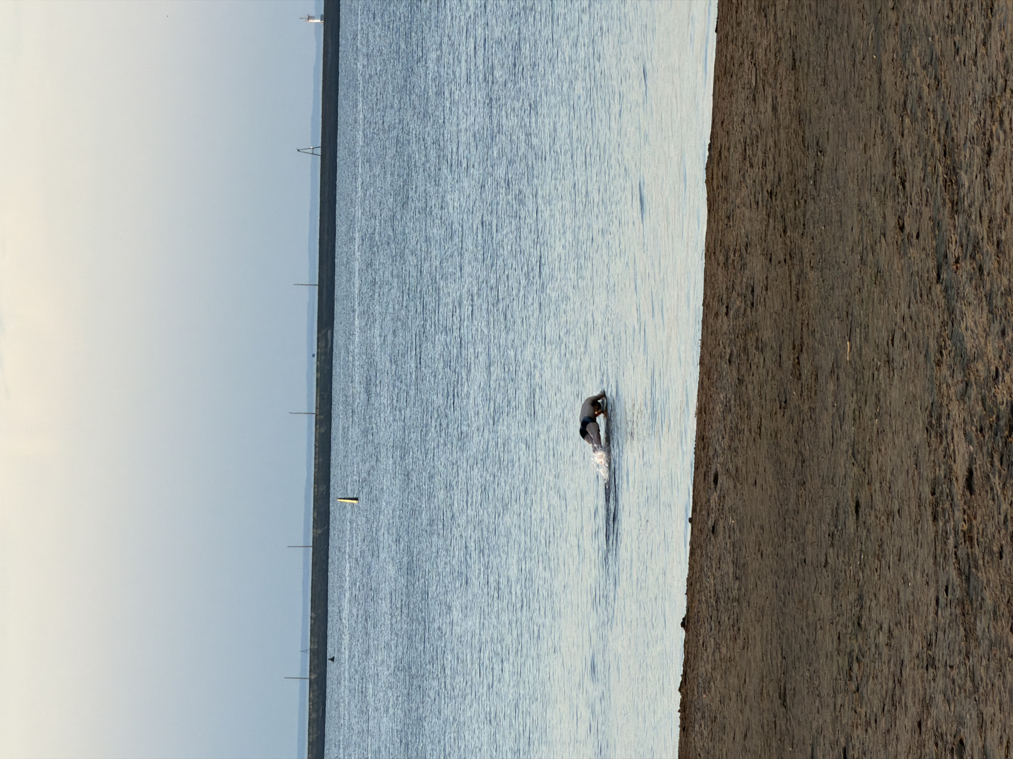 A horizontal eye-level shot depicts a man wading into a body of water. To the left is a section of land with poles visible in the distance, and on the right is a dark brown landmass. The sky is overcast, contributing to a muted color palette. The body of water appears calm with minor ripples, and the man is positioned in the right-center of the frame, appearing to be walking away from the viewer. The scene seems to be taken in Chaikovsky, Russia.