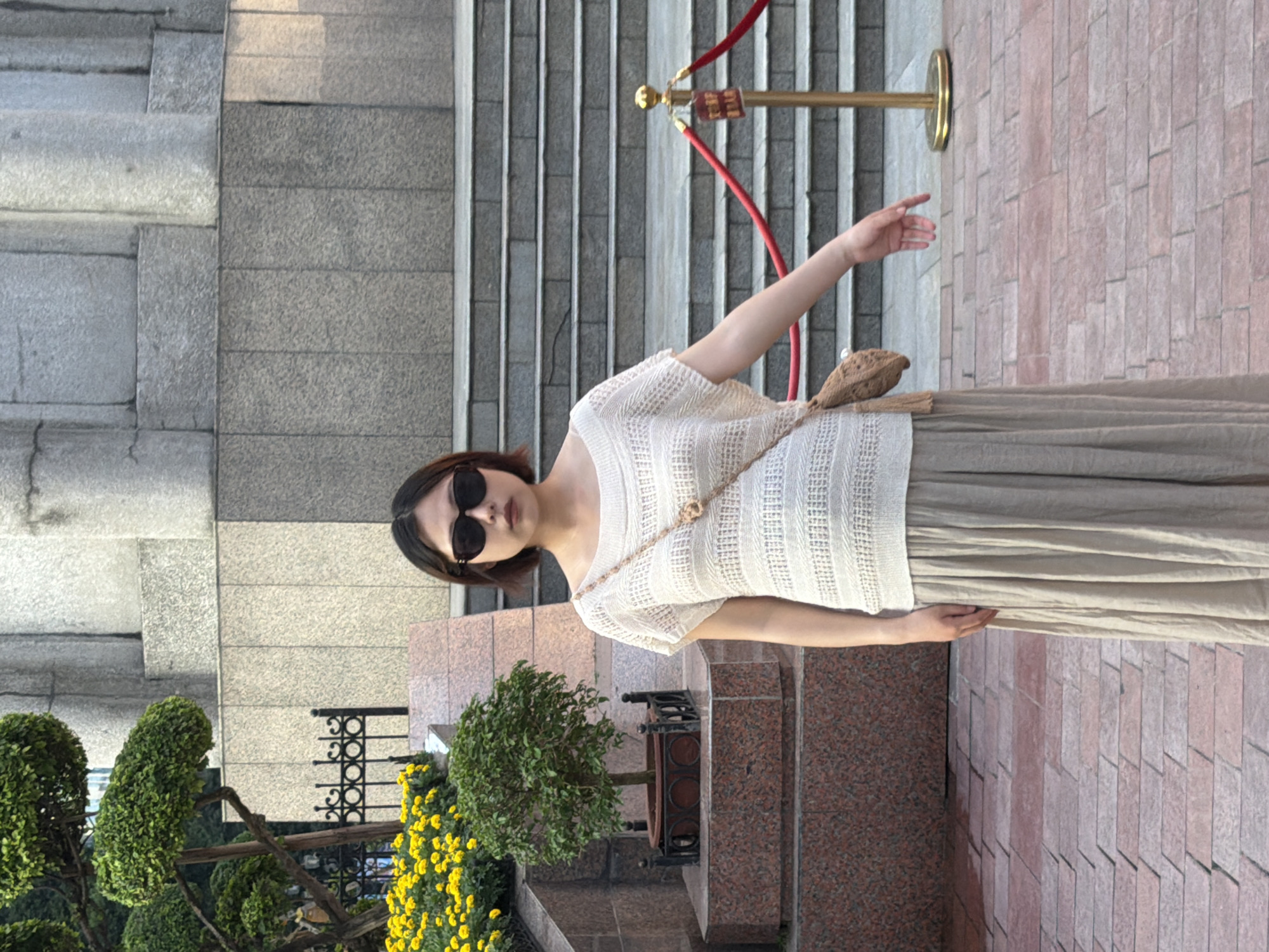 A young woman stands outdoors, posing for a photo. She is wearing a cream-colored, short-sleeved, textured top and a long, flowing beige skirt. A small, woven crossbody bag is slung over her shoulder. She is also wearing dark sunglasses, and her dark hair is cut in a bob. Her left arm is extended, with her hand open, as if reaching out or gesturing. The background shows a grand stone staircase and a paved area with brickwork. A red velvet rope is strung between gold stanchions, suggesting an entrance or restricted area. To the left of the woman, there is lush green topiary and a patch of bright yellow flowers. The lighting suggests it is daytime, possibly late afternoon, with the sun casting shadows. The scene appears to be in a public space, perhaps near a building entrance or public monument. There is no visible text in the image.