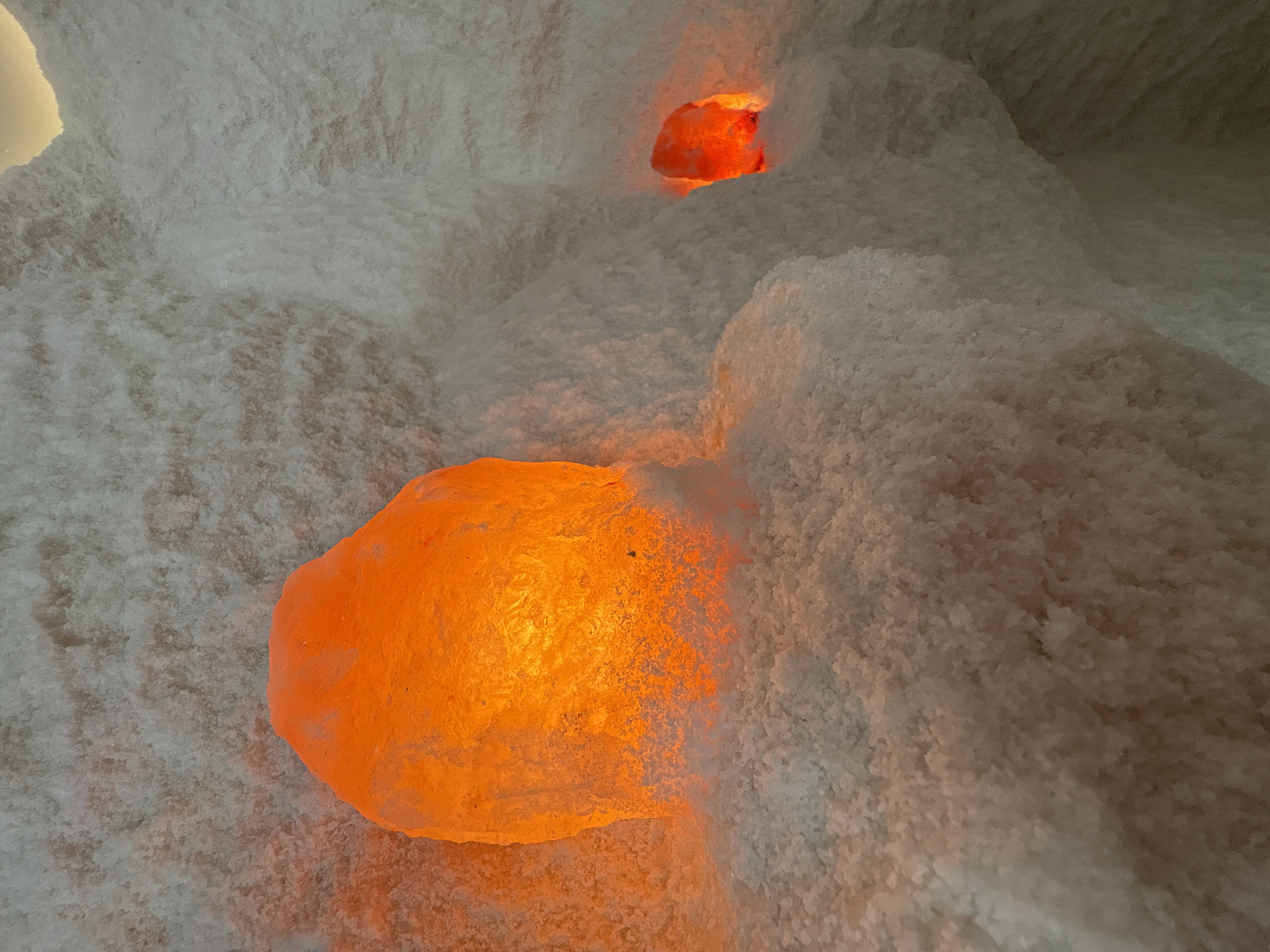The image presents an indoor scene depicting a salt room or salt cave, characterized by rough, crystalline white surfaces. No individuals are visible. The primary subjects are two irregularly shaped, glowing orange objects, which appear to be large Himalayan salt lamps or illuminated salt rocks, embedded within the textured white material. One larger glowing orange object occupies the lower left foreground, emitting a vibrant, warm light that casts an amber glow onto the surrounding salt. A second, smaller orange light source is visible further back and higher in the frame, similarly nestled within the white salt formations.
The environment is composed of a coarse, granular white substance, likely salt crystals, which form organic, uneven mounds and crevices, creating a natural, cave-like aesthetic. The lighting is low and atmospheric, originating solely from the warm orange lamps. This artificial illumination highlights the textures of the salt and creates a tranquil and relaxing ambiance. The scene suggests a setting for wellness or therapy, such as a halotherapy room or a spa. No text is visible in the image. Given the context, this environment is situated in Anapa, Russia. The time of day and weather are not relevant due to the indoor, artificially lit setting.