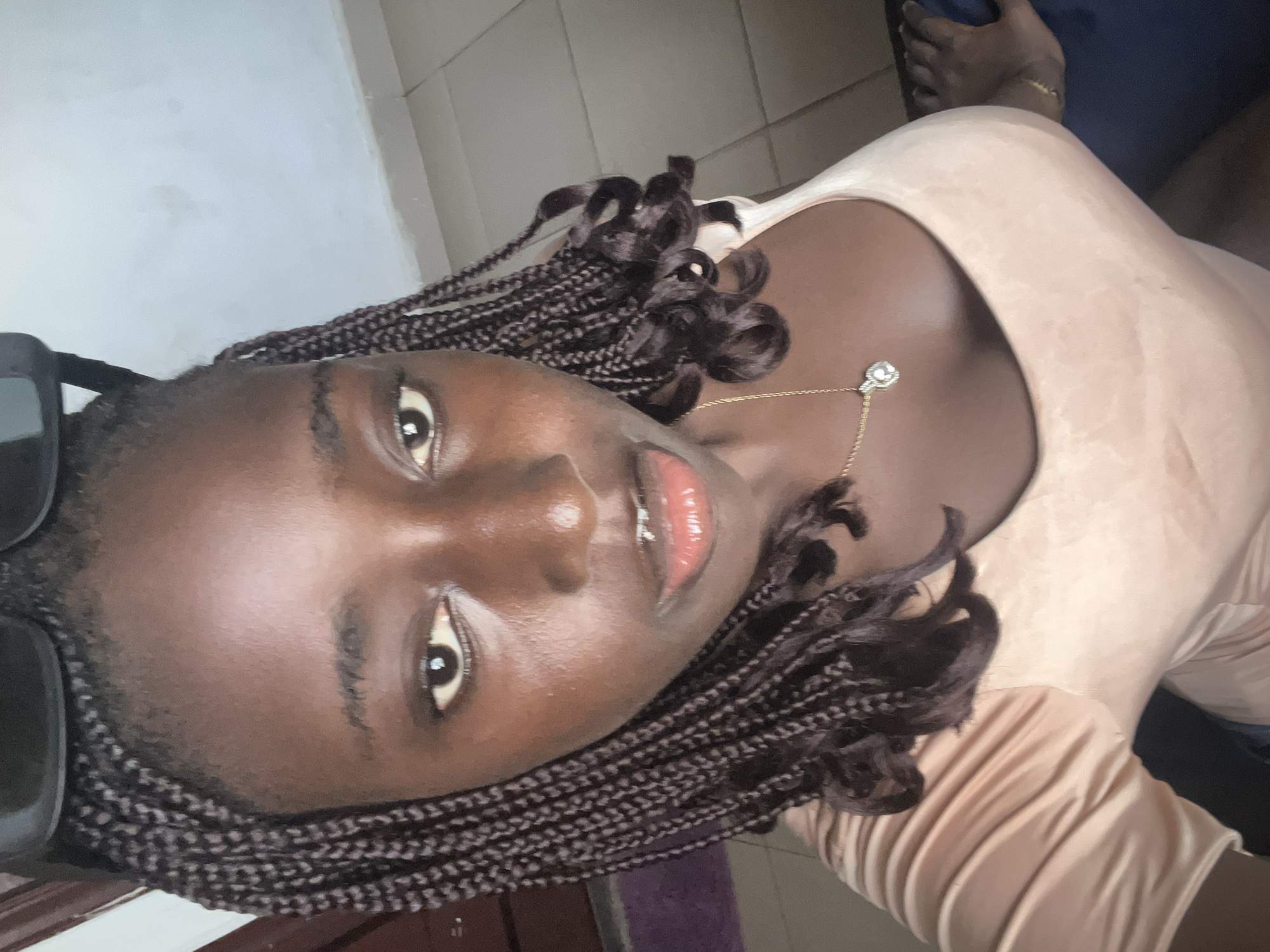 A young woman with dark skin and braided hair is photographed indoors. She is wearing a light-colored top and a necklace with a pendant. A pair of sunglasses rests on her head. The background appears to be a tiled floor and part of a wall. The location is not discernible from the image.