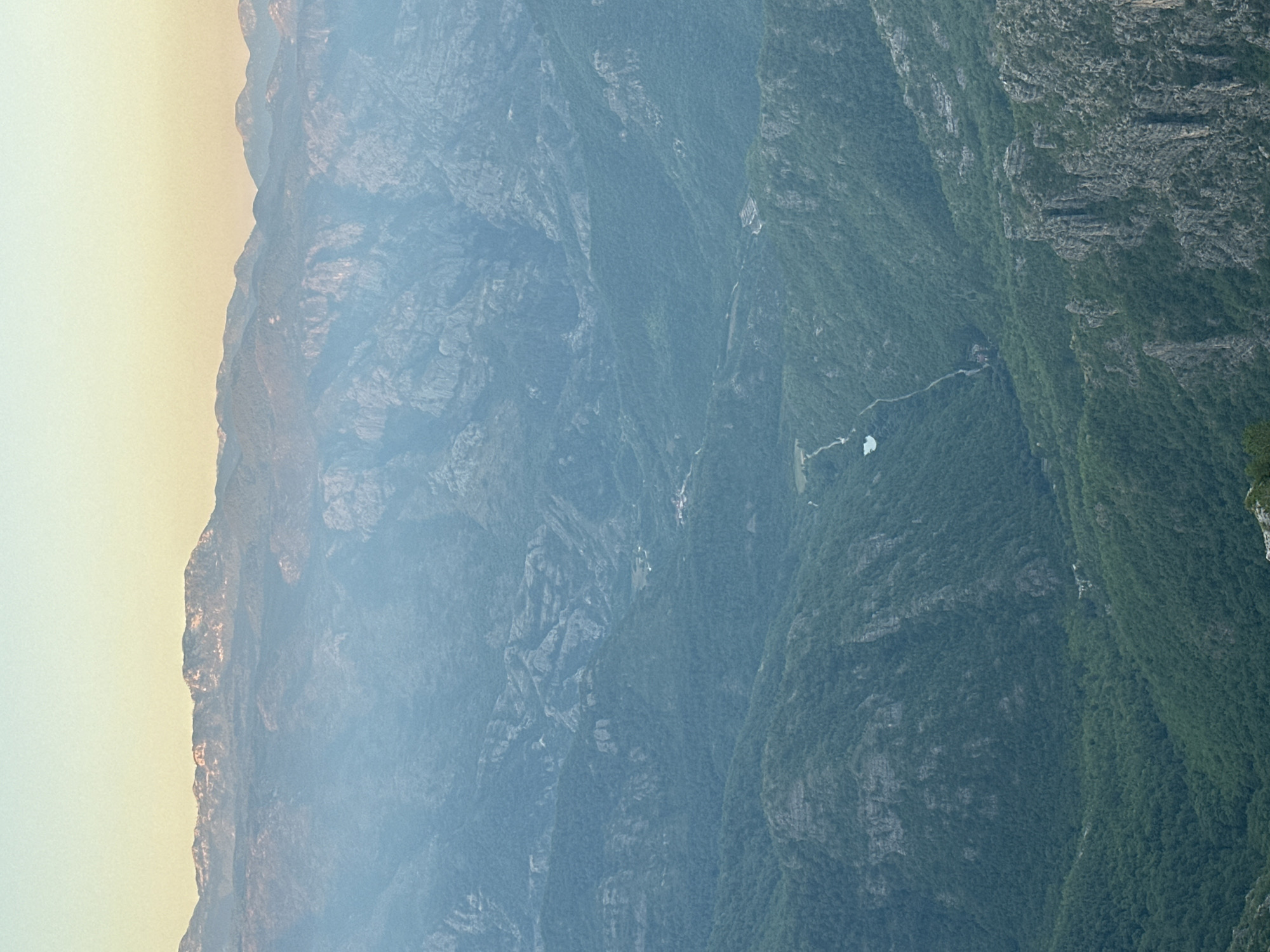 This is an aerial landscape photograph showing a vast mountainous region. The scene is dominated by layers of hazy green mountains receding into the distance, with the sky transitioning from a light yellow near the horizon to a pale blue at the top of the frame. In the foreground, rough, rocky terrain with sparse vegetation is visible. A winding valley cuts through the middle ground, with a small body of water and a few scattered buildings nestled within the dense green forest covering the slopes. The mountains in the background are jagged and appear to be covered in a lighter green foliage or possibly sparse vegetation, with some areas showing lighter rock formations. The overall impression is one of a remote, rugged, and undeveloped natural environment. There are no people or specific actions depicted in the image. The image does not provide any visual cues to suggest it is located in Aranđelovo, Bosnia and Herzegovina.