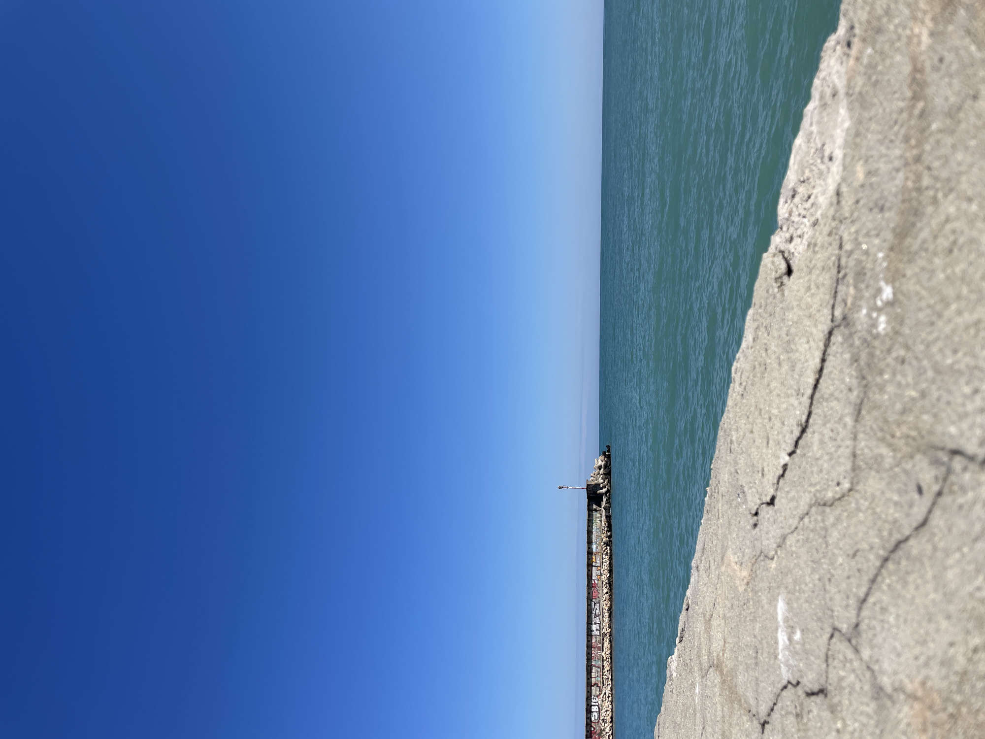 The image presents a scene from Varna, Bulgaria, featuring the sea and a pier or breakwater. A clear blue sky occupies the left side of the frame, contrasting with the blue-green hue of the water on the right. A concrete structure, presumably part of the pier, is visible in the foreground, exhibiting cracks and weathered surfaces. Further along the pier, towards the center of the image, a light pole and a cluster of large rocks or concrete blocks are positioned. The pier appears to have some graffiti on it. In the upper right corner of the pier, two dark silhouettes might be people standing or sitting. The photograph is taken at an oblique angle, offering a perspective that emphasizes the horizon line and the expanse of the sky and sea.