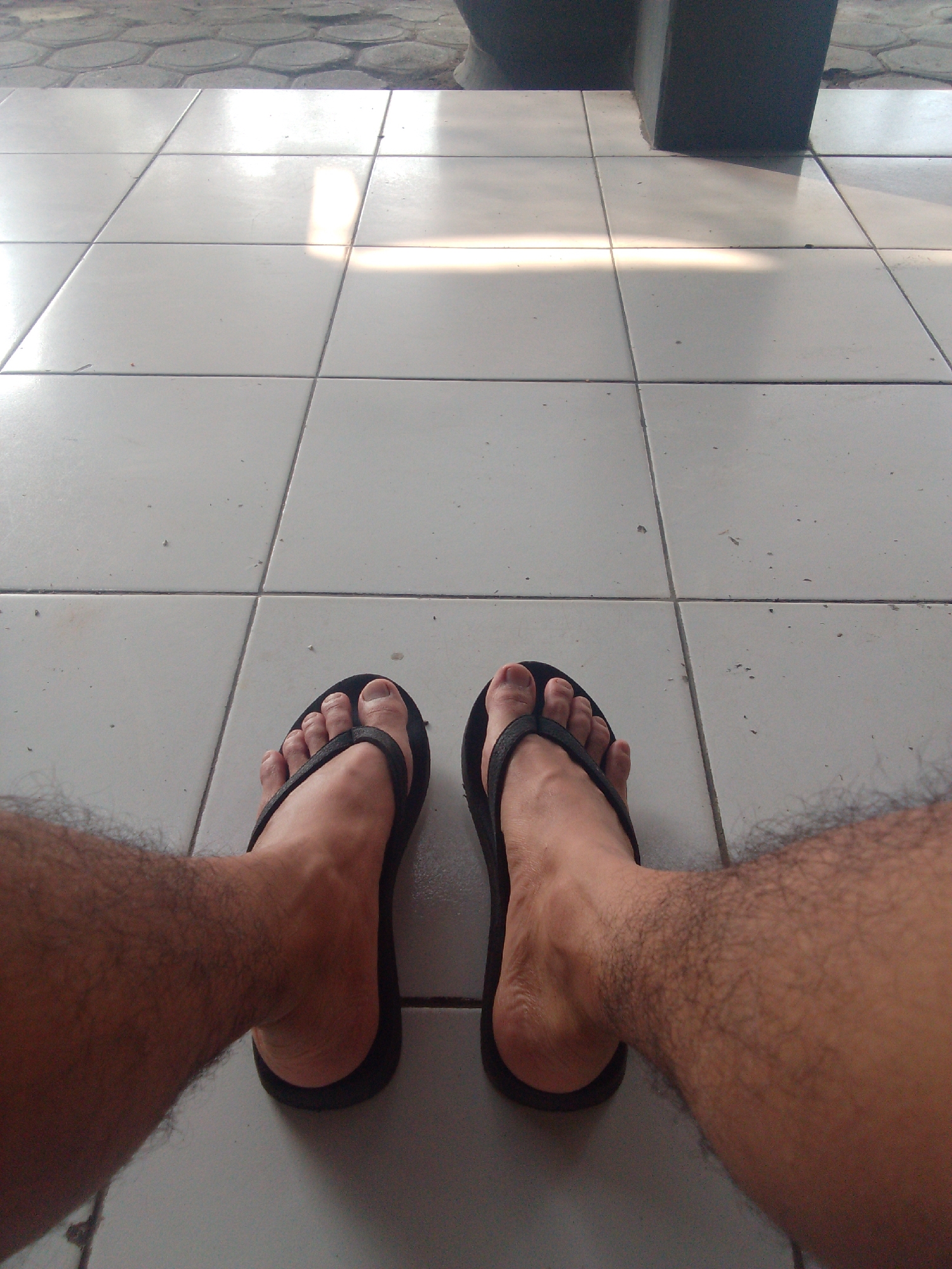 The image shows a first-person perspective of a person's legs and feet wearing black flip-flops. The person is sitting on a tiled floor, with hexagonal paving stones visible in the background. The lighting suggests it is daytime, with sunlight casting shadows across the tiles. The scene appears to be an outdoor or semi-outdoor area, possibly a patio or porch. The location is identified as the city of Trenggalek, Indonesia. The image contains no other subjects or objects of note.