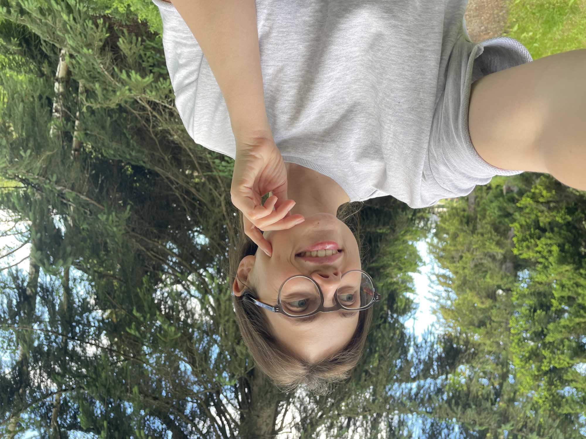A young woman wearing a grey t-shirt and round glasses is taking a selfie outdoors. She is smiling and her right hand is touching her chin. She is positioned in front of a backdrop of lush green trees, some of which are coniferous. The background suggests a natural setting, possibly a park or forest. The lighting indicates it is daytime, with sunlight filtering through the trees. The scene appears to be peaceful and serene.
