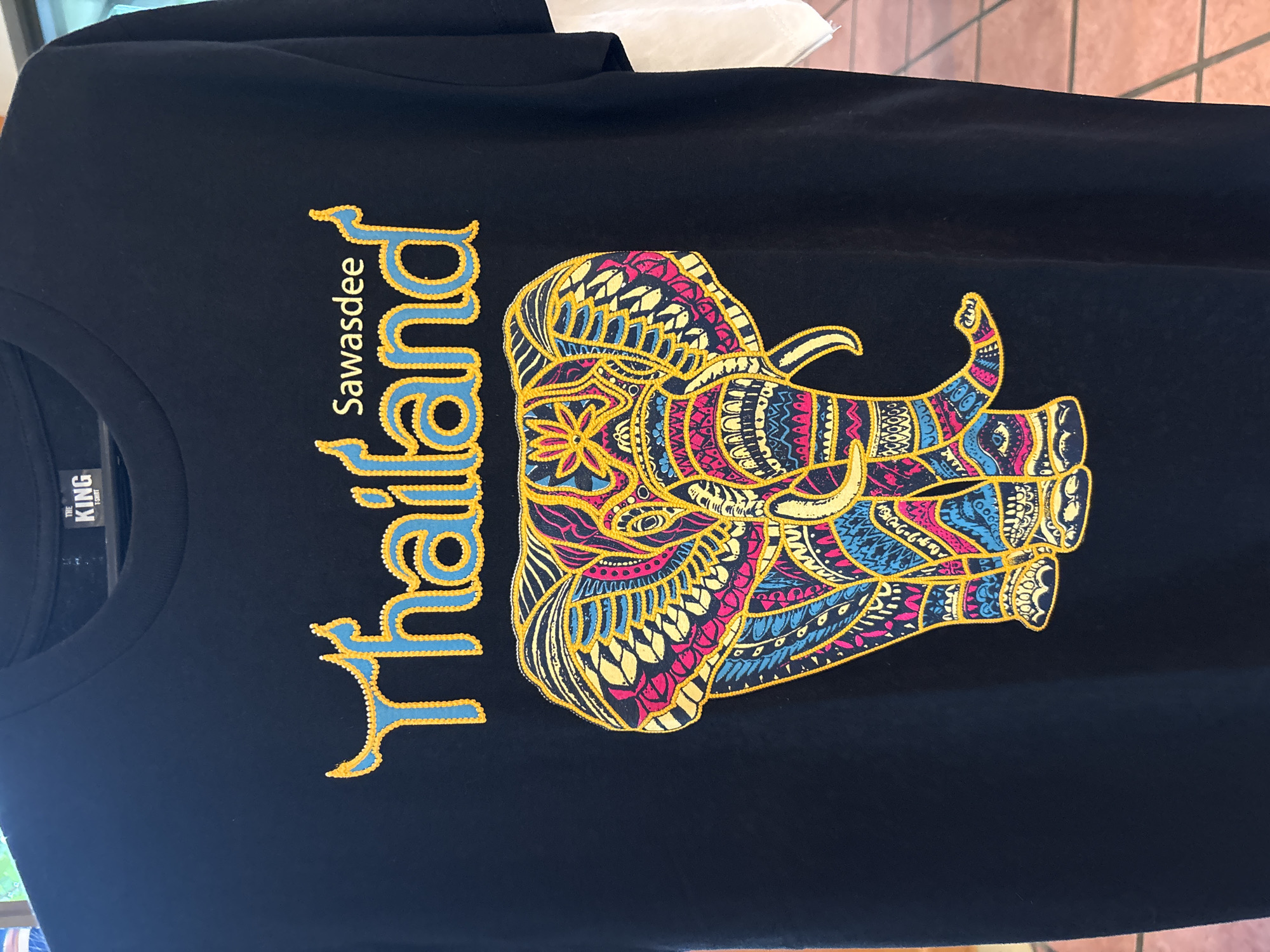 A black t-shirt with a colorful elephant design is displayed. The t-shirt has a crew neck and short sleeves. The graphic on the t-shirt features the word "Thailand" in blue and yellow lettering, with "Sawasdee" written above it. Below the text is an intricately patterned elephant in shades of pink, blue, yellow, and black. The t-shirt is hanging on a hanger. The background appears to be an indoor setting with tiled flooring and a hint of a wall. The location is identified as Chalong, Thailand.