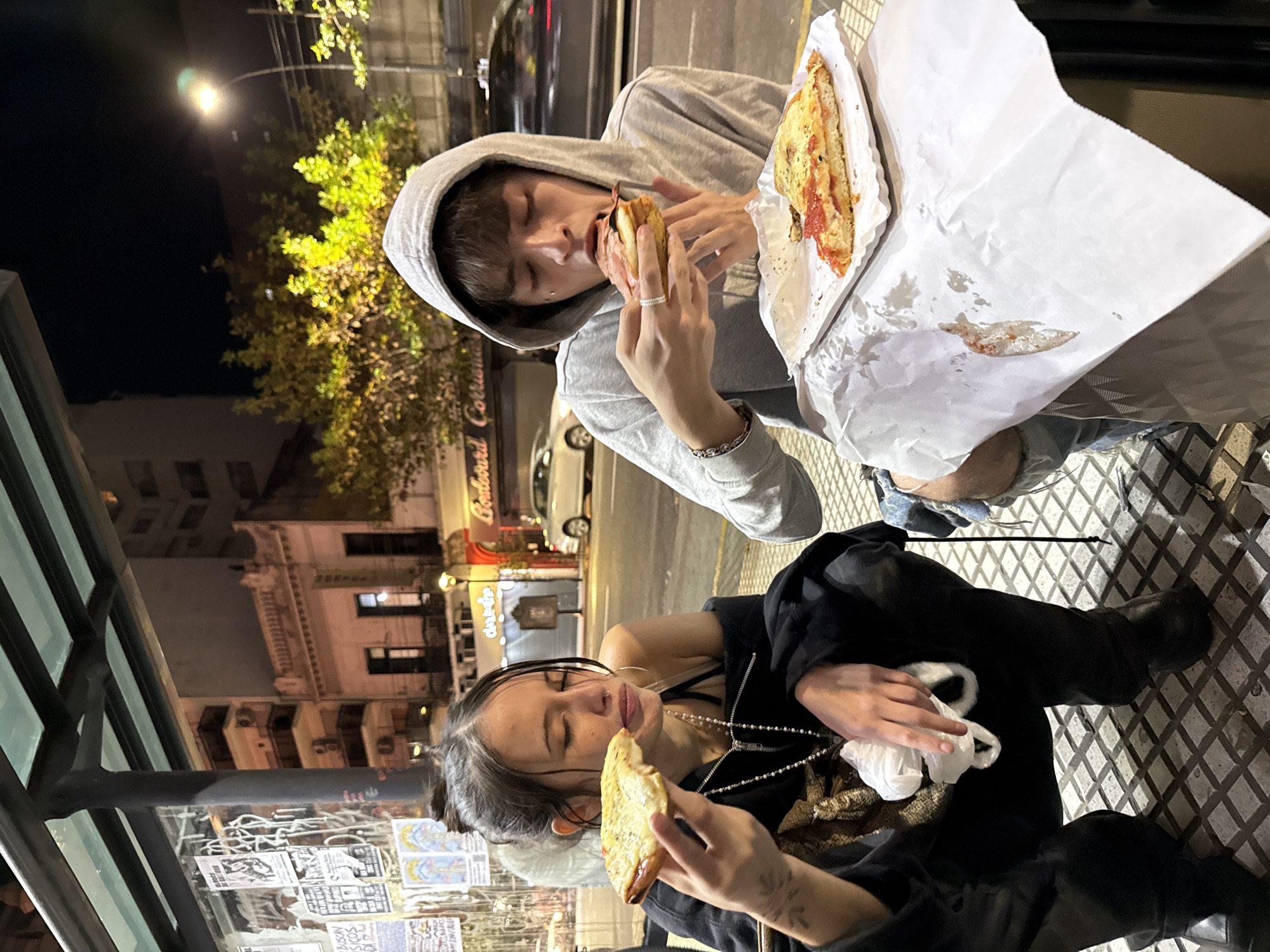 The image captures two individuals eating pizza on a street in Buenos Aires, Argentina. The lighting suggests it is night.
A person wearing a gray hooded sweatshirt is seated and holds a slice of pizza, mid-bite, in their hands. There is a larger pizza slice alongside them on a white paper. They wear a ring on their finger and a bracelet.
The second person is seated below and to the left of the first. They are wearing a black shirt or jacket, jewelry, and dark-colored pants. They also hold a slice of pizza.
They appear to be sitting at or near a bus shelter, indicated by the shelter's metal frame on the left of the frame, and a series of posters on the wall of the shelter. The background reveals a street scene with buildings, signage, and street lighting indicating an urban environment.