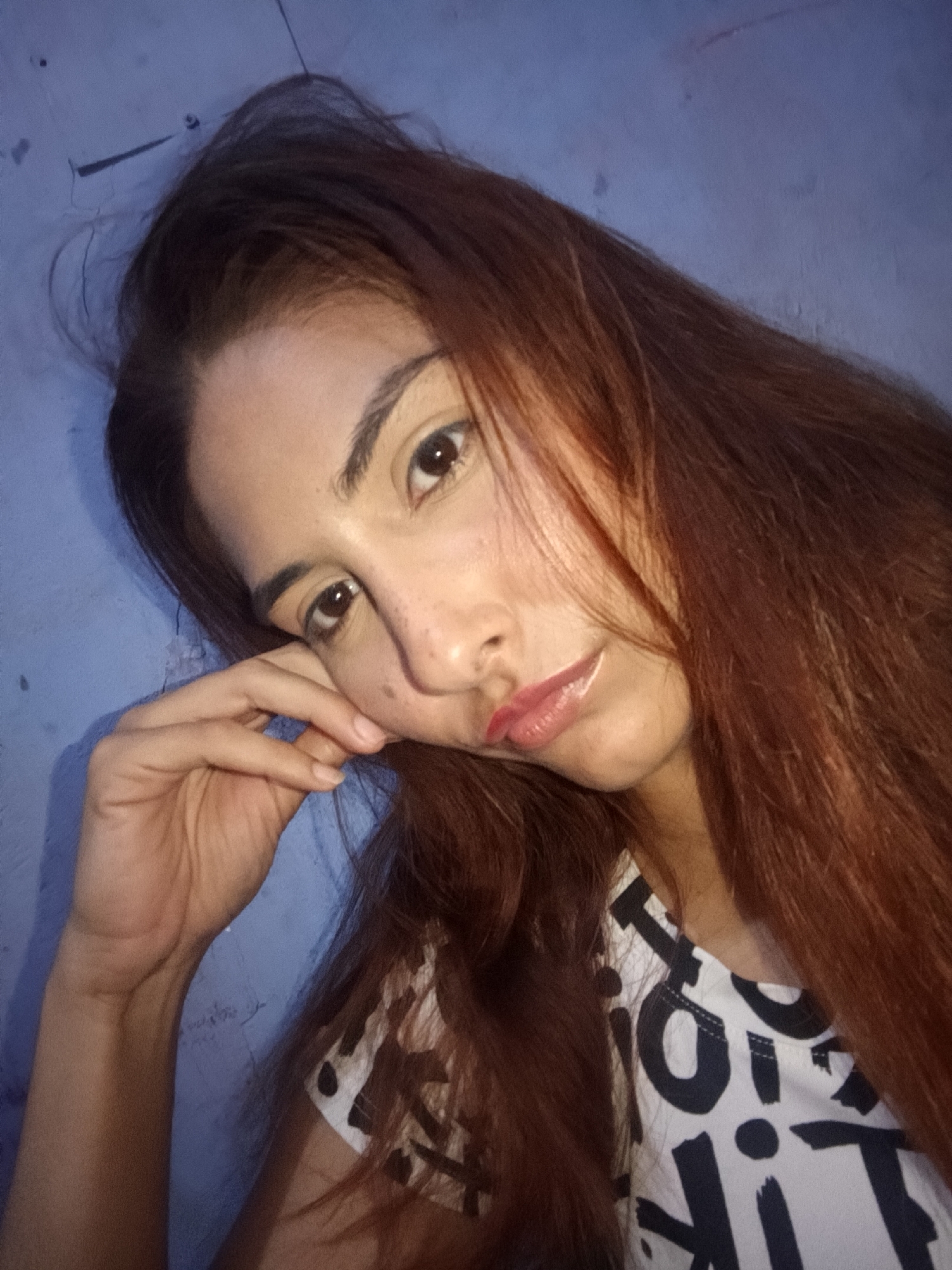 A young woman with long, reddish-brown hair is leaning her head on her hand, with a melancholic expression. She is wearing a white t-shirt with black lettering. The background appears to be a blue wall. There is no indication of a location in Juan Griego, Venezuela.