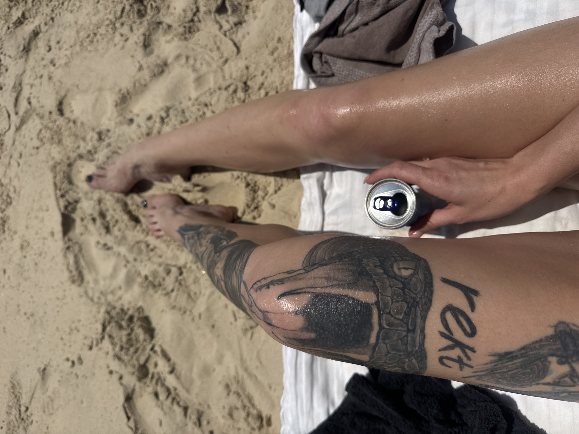 A woman is lying on a beach towel on a sandy beach. The photograph is taken from an overhead perspective, focusing on her legs, which are extended towards the viewer. Her left leg has tattoos, including a snake design and the word "text". She is holding an open can in her right hand. The location is a beach, characterized by the sand. There is no further information to confirm if the location is in Bat Yam, Israel.