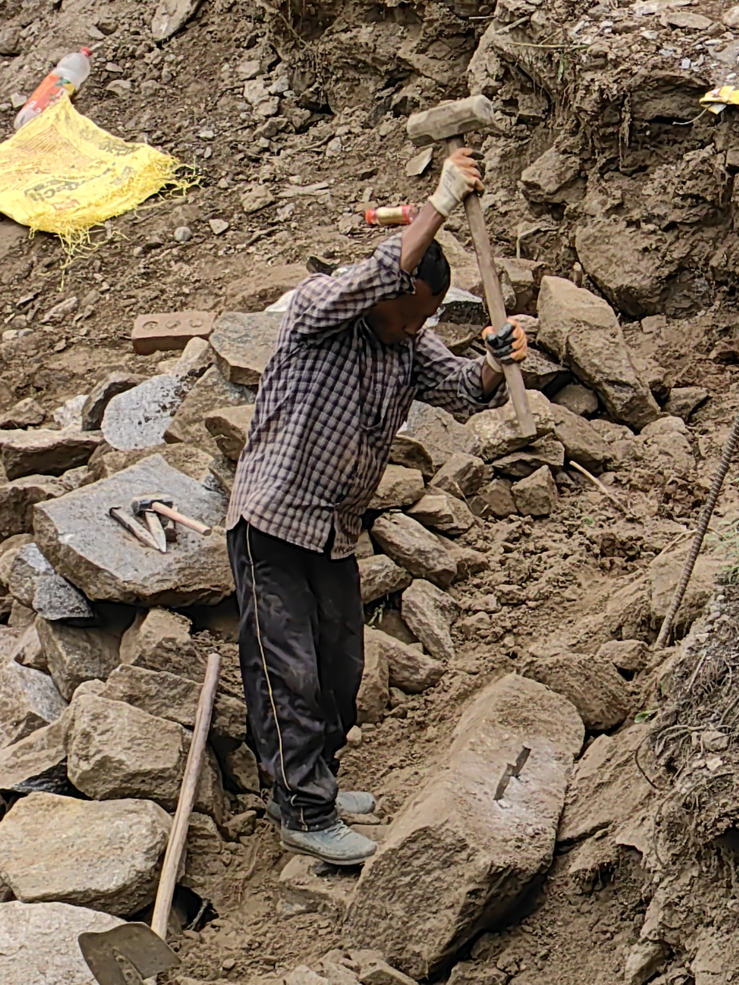 A person is engaged in stone breaking or shaping in a rocky, outdoor environment, possibly a construction site or a quarry, in Vashisht, India. The individual is wearing a checkered shirt, dark pants with a yellow stripe, and light-colored gloves. They are using a large sledgehammer to strike a substantial rock.
Several other tools are visible: a smaller hammer and chisels are laid on a flat stone to the left of the main subject. A shovel and a wooden pole are also present in the foreground. Scattered around are numerous rocks of varying sizes, some appearing rough and unworked, others possibly in the process of being shaped. A yellow bag, possibly containing cement or construction material, lies on the ground to the upper left, along with a partially filled plastic bottle.
The scene appears to be outdoors, with natural lighting suggesting daytime. The terrain is uneven and dusty, composed of earth and rocks. The activity is labor-intensive, as indicated by the use of the sledgehammer. No specific time of day or weather conditions can be definitively determined, though the lighting suggests it is not dawn or dusk. There are no discernible emotions from the subject, as their face is partially obscured. There is no visible text in the image, apart from what might be on the yellow bag, which is not legible.